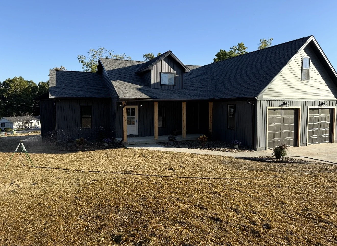 A modern two-story house with a dark exterior, multiple gabled roofs, and an attached three-car garage. The house is under sunny weather with a clear blue sky, and the front yard has a patch of yellowish grass with a few small potted plants near the porch.