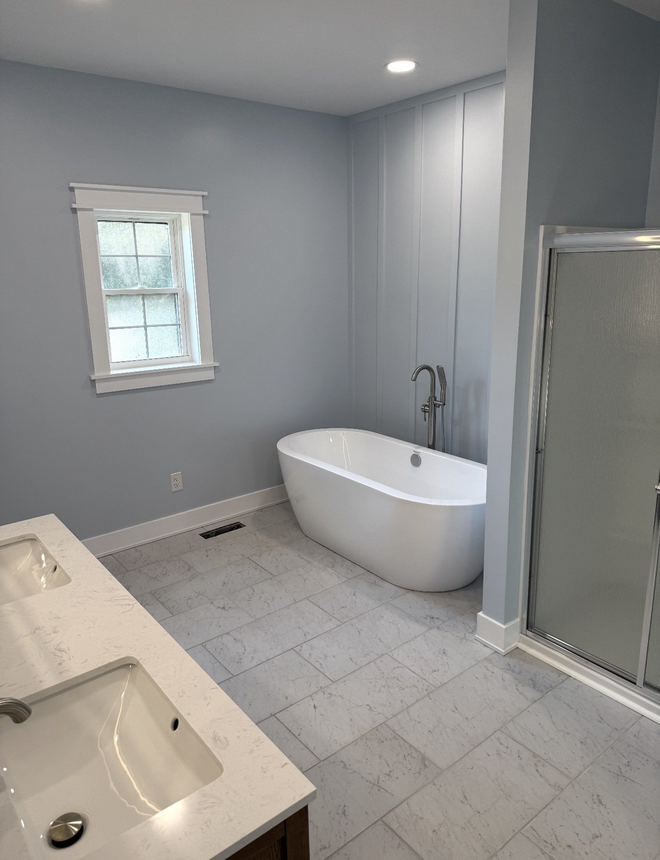 Bathroom with light blue walls, white bathtub, double sink vanity, frosted glass shower, and a small window.