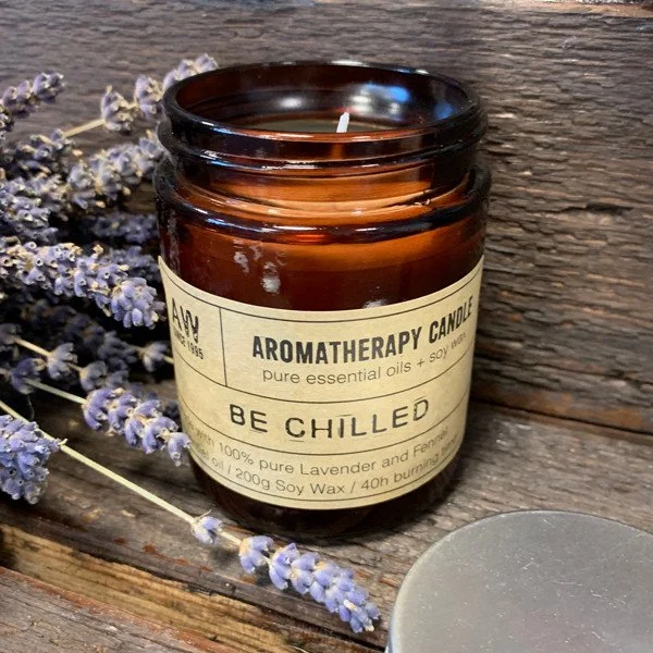Vela aromática en frasco de vidrio marrón con etiqueta que dice 'Aromatherapy Candle, pure essential oils + soy wax, be chilled' y flores de lavanda a su lado, sobre madera.