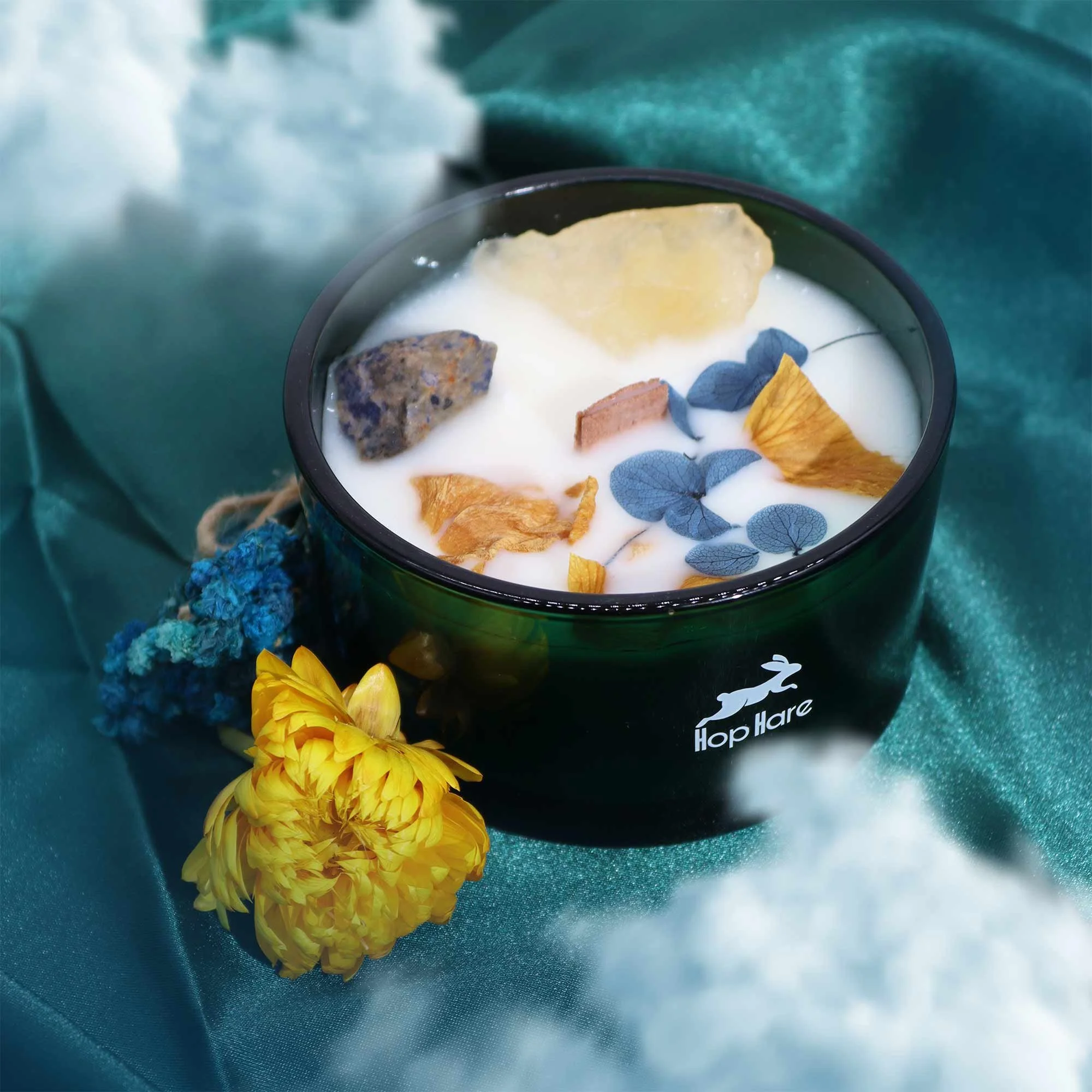 Vela aromática blanca con piedras decorativas y hojas secas en un recipiente negro con la marca Hop Hare, rodeada de flores amarillas y azules, sobre tela azul con nubes de algodón en el fondo.