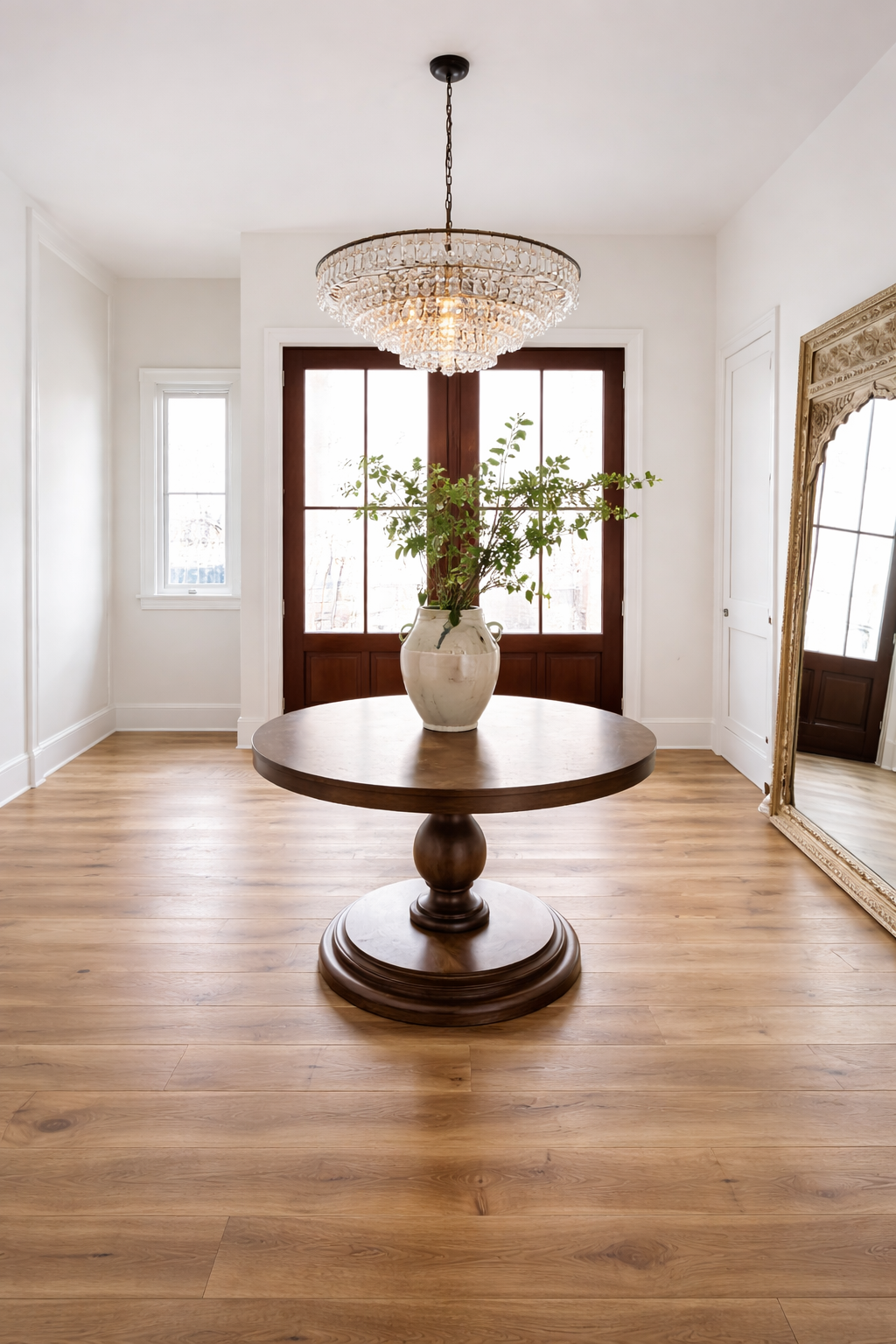 A round wooden table with a white ceramic vase containing green leafy branches, in a bright room with hardwood floors, large window doors, a chandelier hanging from the ceiling, and a large ornate mirror on the right wall.