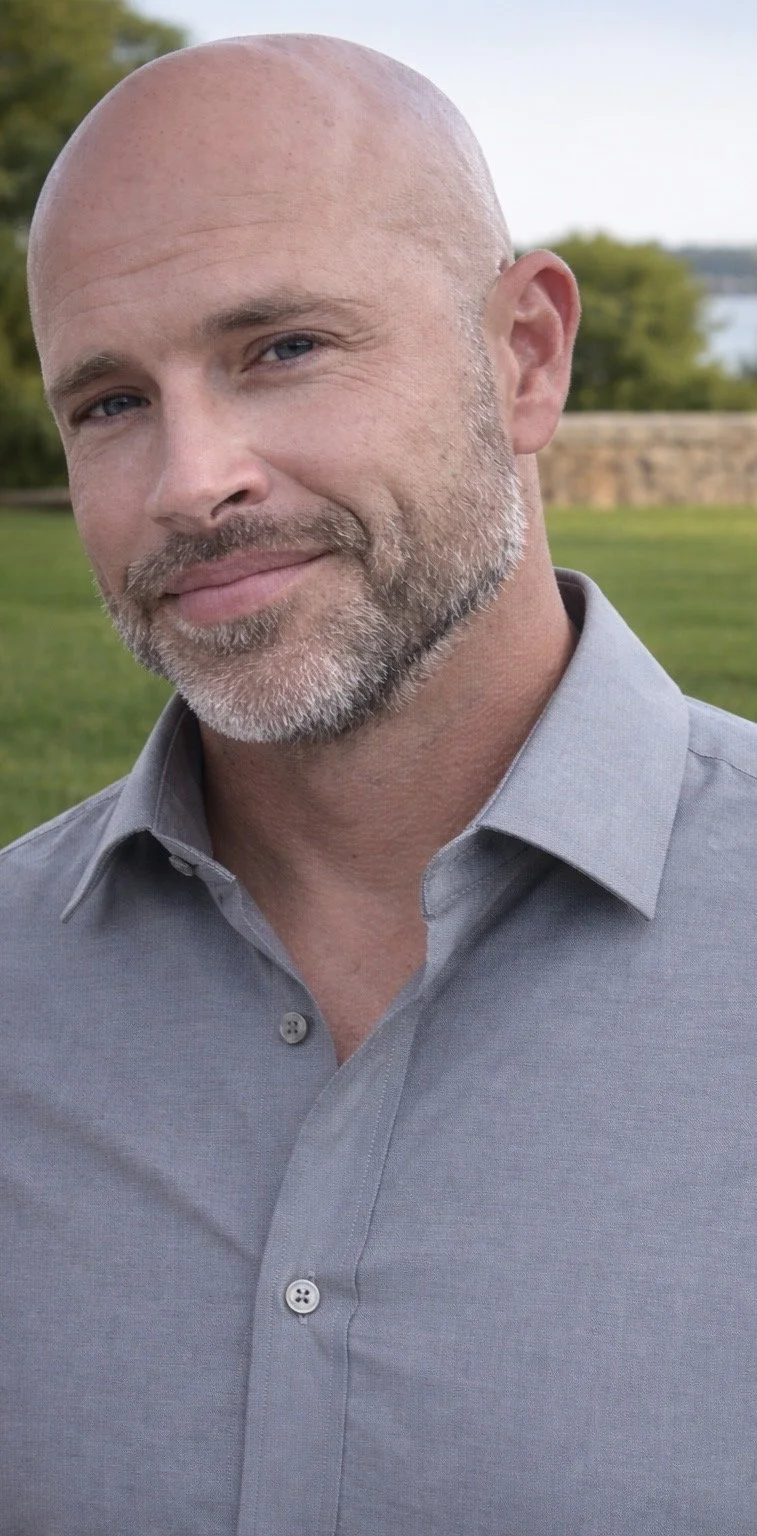 A middle-aged bald man with a beard smiling outdoors, wearing a light gray button-up shirt.