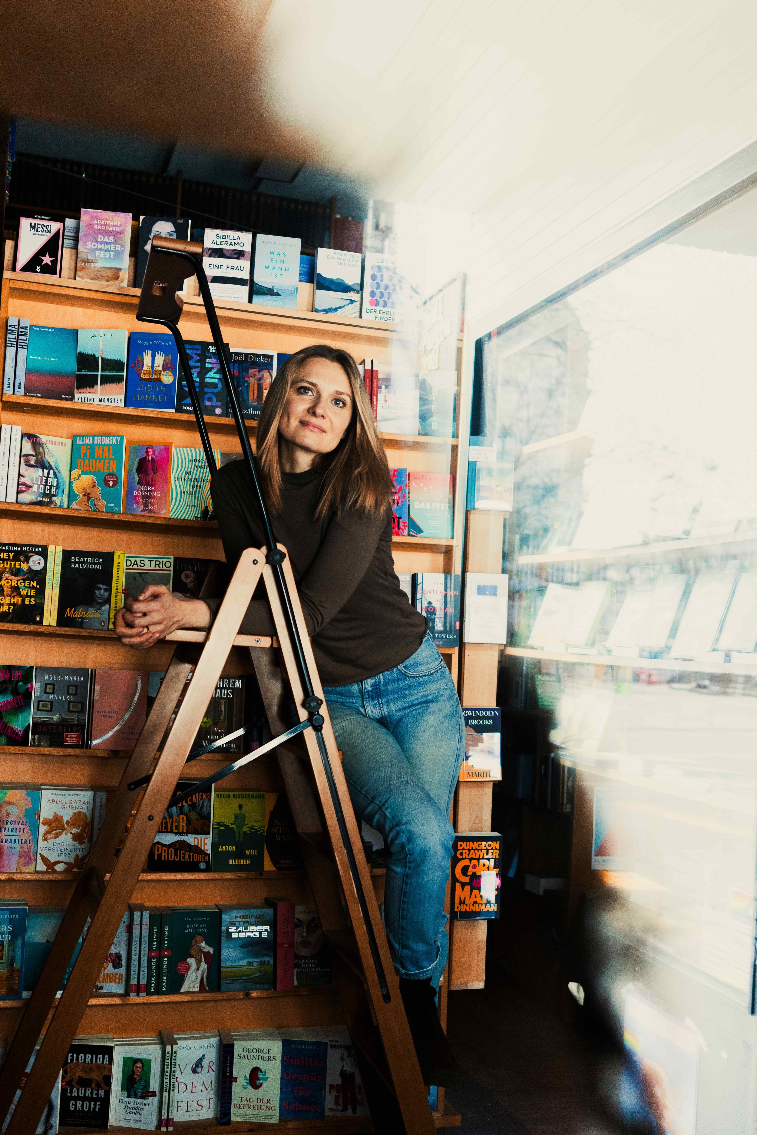 Eine Frau lehnt an einem Bücherregal in einer Buchhandlung oder Bibliothek, mit einer Leiter vor sich, um die oberen Regale zu erreichen. Der Raum ist hell erleuchtet, mit großen Fenstern auf der rechten Seite, die natürliches Licht hereinlassen. Das Regal ist voll mit bunten Büchern.
