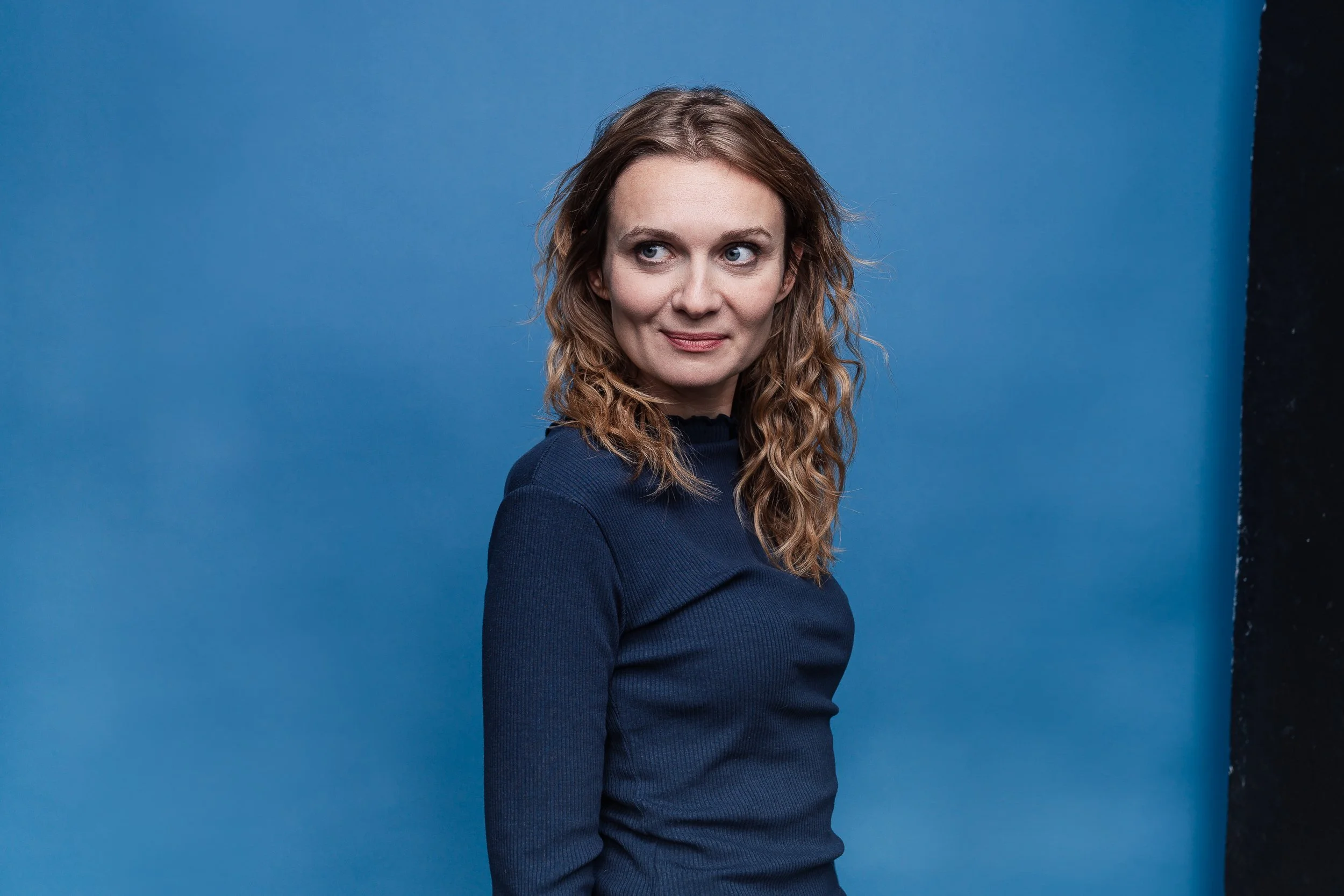 Eine Frau mit lockigem braunem Haar trägt einen dunkelblauen Pullover vor einem blauen Hintergrund.