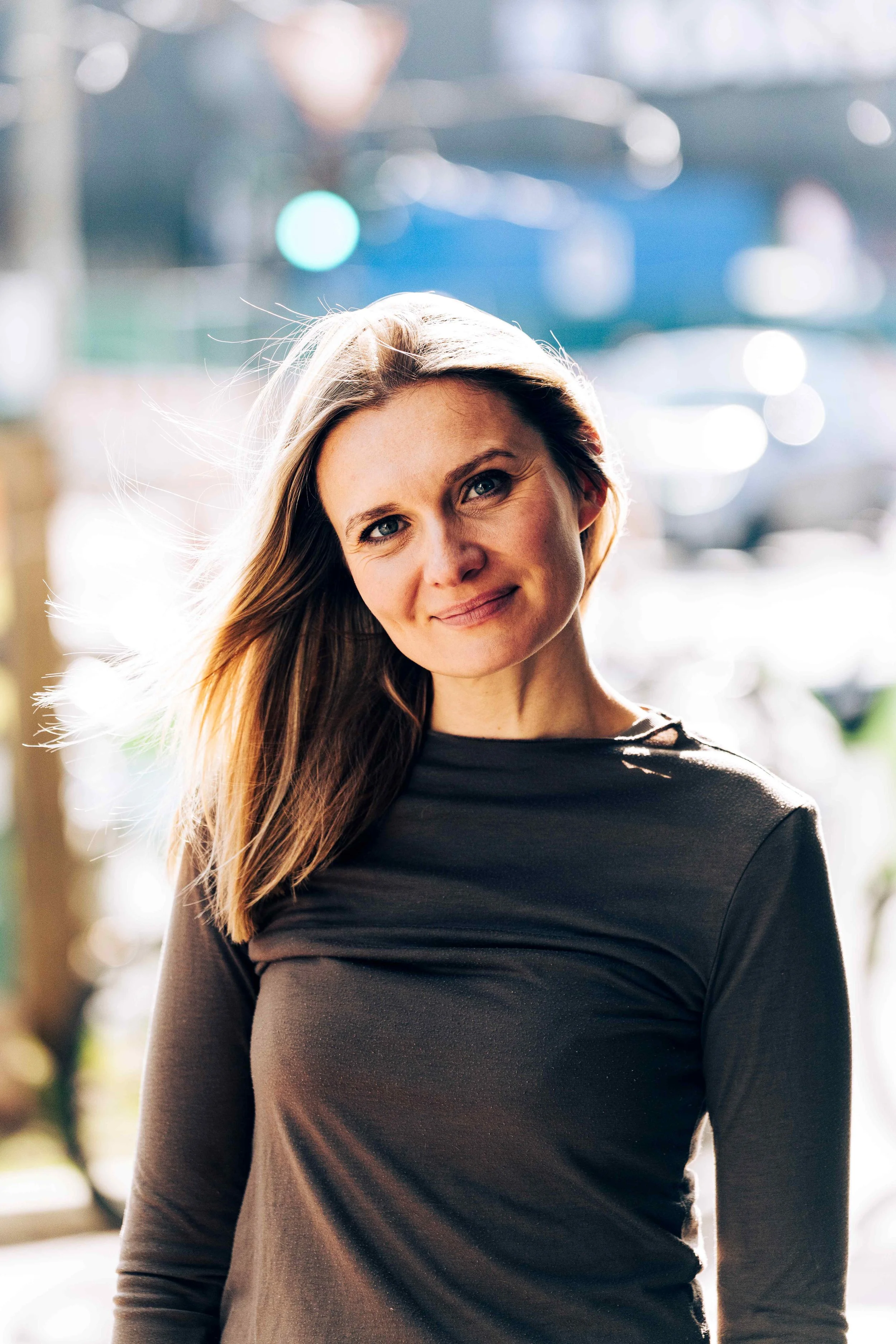 Junge Frau mit langem blondem Haar, die leicht lächelt und in der Sonne auf einer Straße steht.