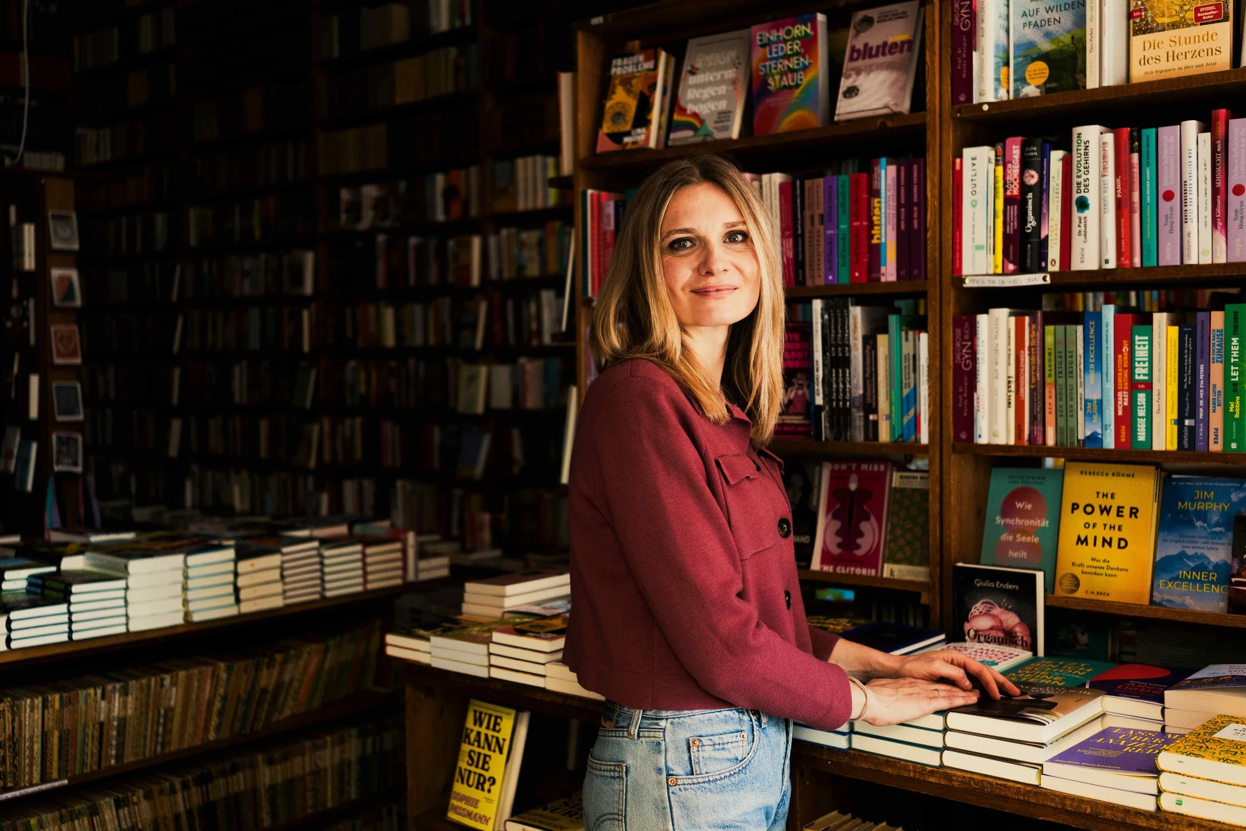 Eine junge Frau steht in einer Buchhandlung, umgeben von Regalen voller Bücher, und blättert in einem Buch auf einem Tisch.