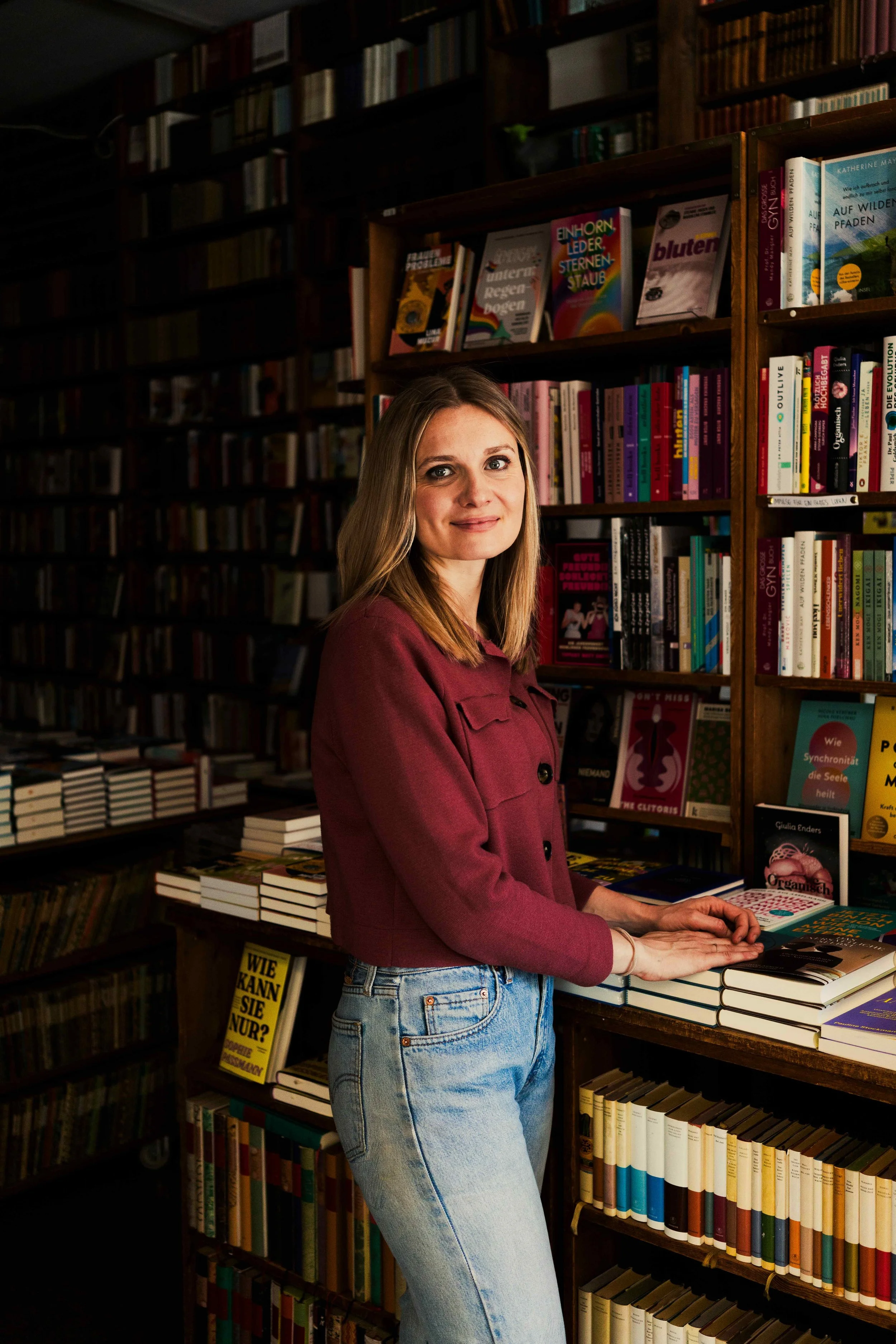 Junge Frau mit blonden Haaren in einem Bücherladen, trägt eine rot-bordeauxfarbene Jacke und blue Jeans, steht vor Büchernregalen und liest ein Buch.