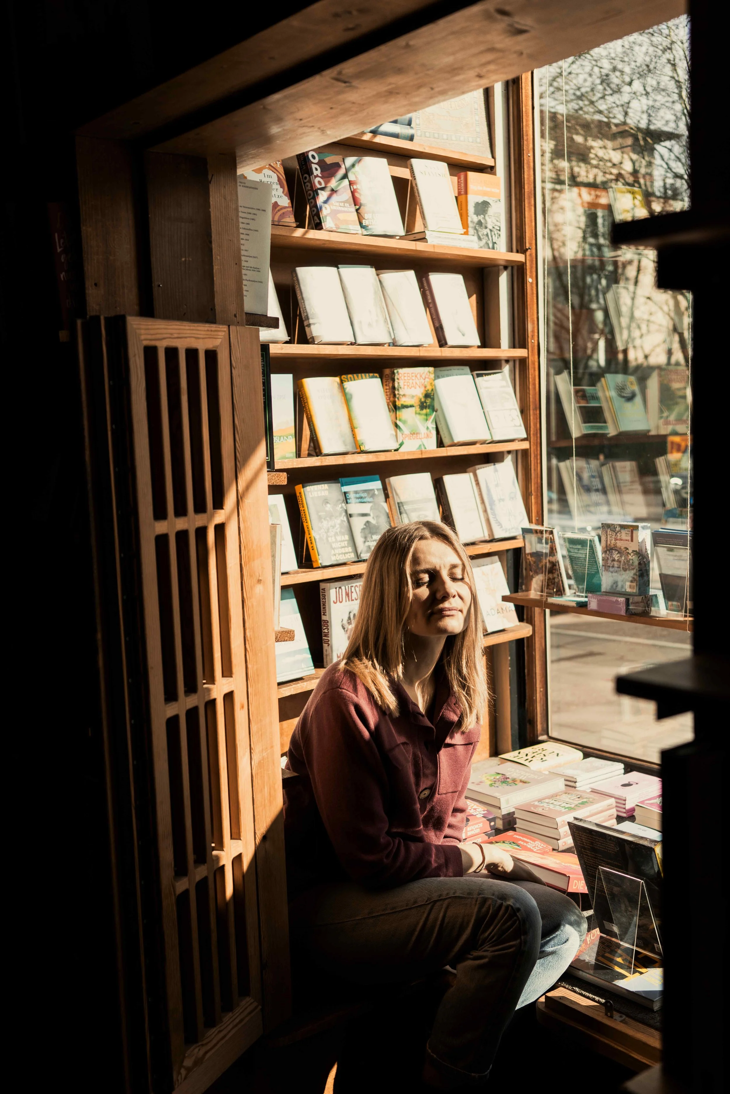 Junge Frau sitzt in einer Buchhandlung am Fenster, Sonnenlicht beleuchtet ihr Gesicht und die Bücherregale im Hintergrund.