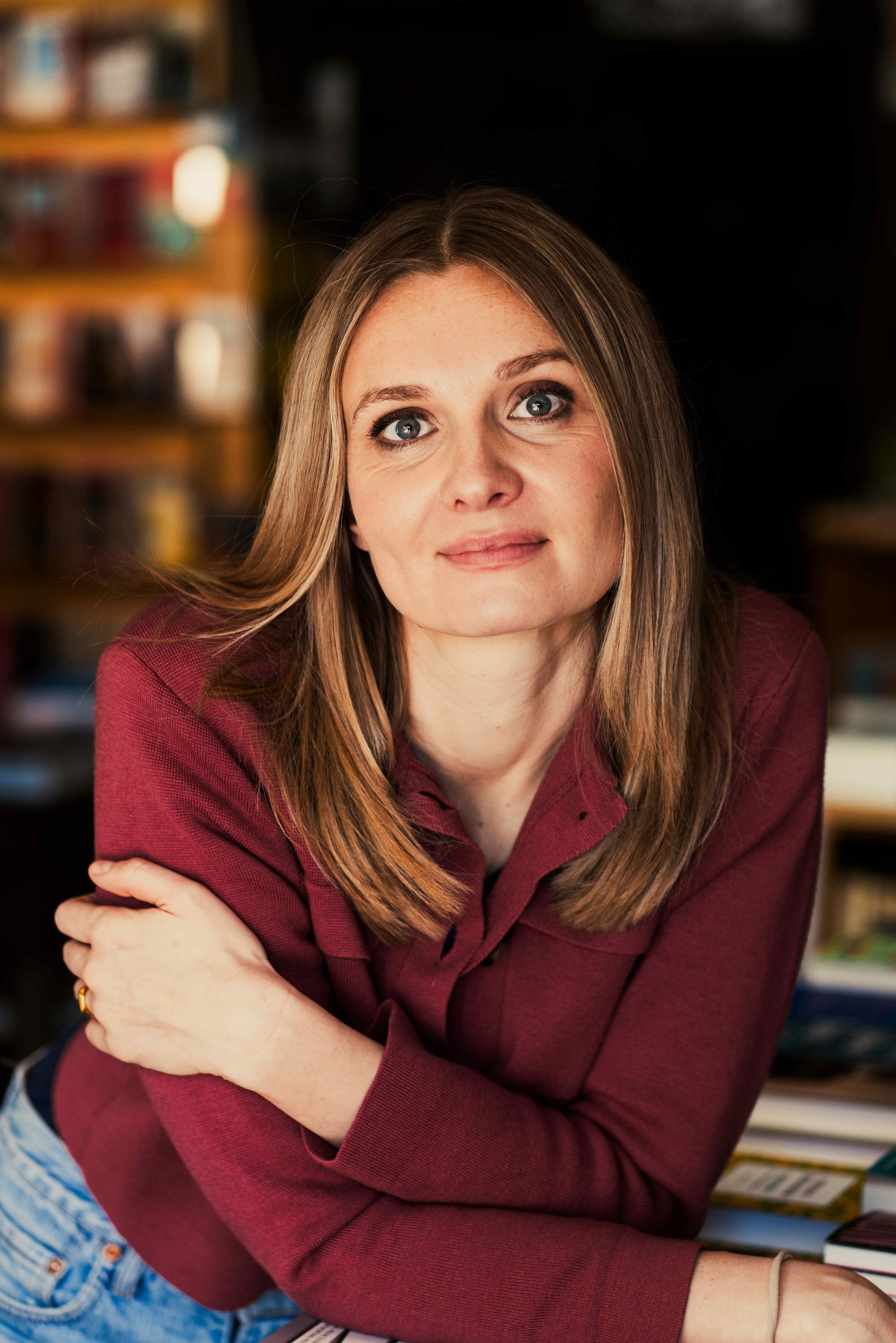 Eine junge Frau mit langem, blondem Haar, blauen Augen und einem hellen Teint sitzt in einem Raum mit Bücherregalen im Hintergrund. Sie trägt ein rotes Hemd und schaut direkt in die Kamera, mit einem leichten Lächeln.