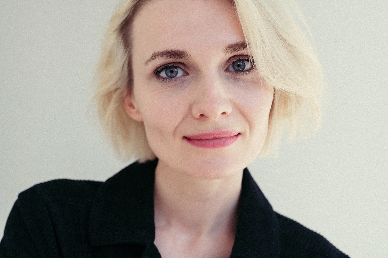 Nahaufnahme eines jungen Mädchens mit blonder, kurzer Frisur und blauen Augen, trägt ein schwarzes Jackett, vor hellem, neutralem Hintergrund.