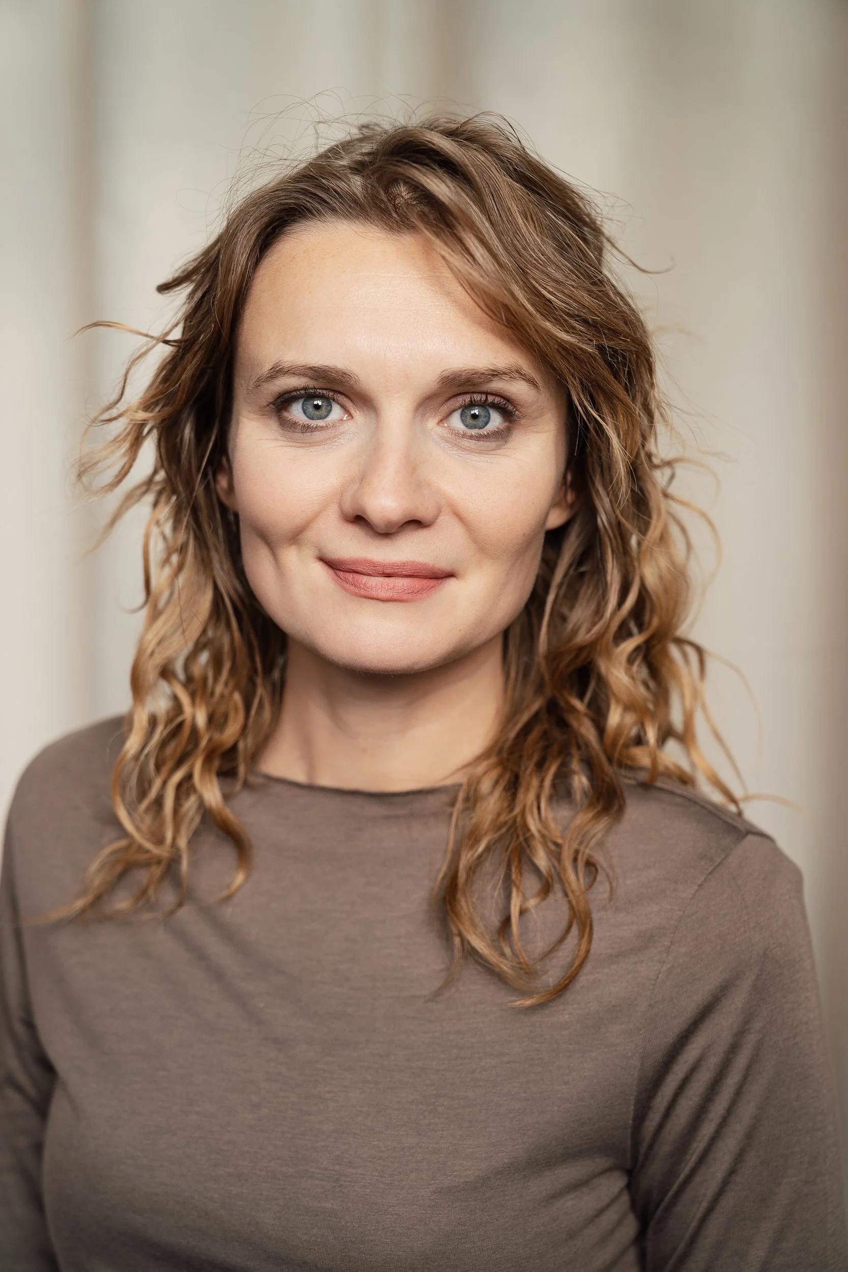 Nahaufnahme einer Frau mit lockigem, braunem Haar und blauen Augen, die ein neutrales Lächeln zeigt, vor einem neutralen Hintergrund.