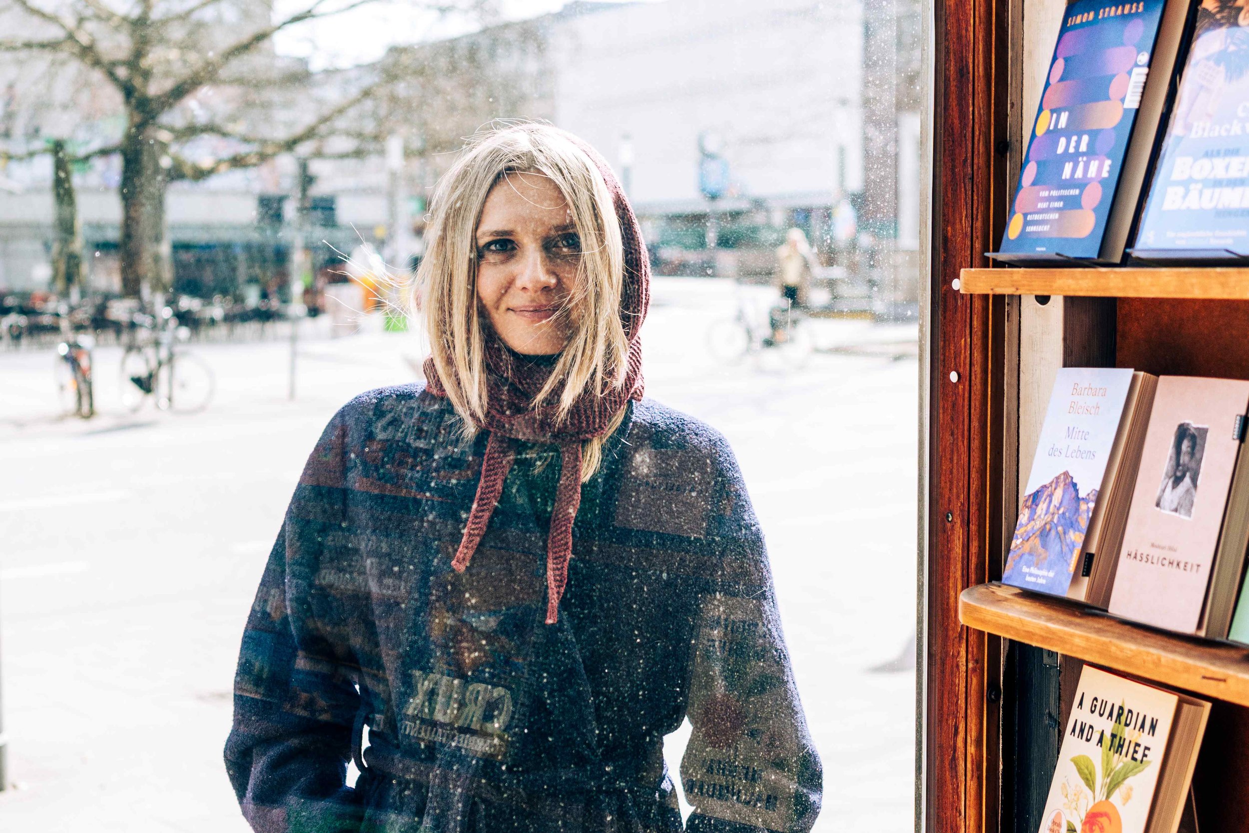 Frau blickt durch ein Schaufenster in eine belebte Straße mit Fahrradständern und Bäumen, neben ihr ein Regal mit Büchern in einem Laden.