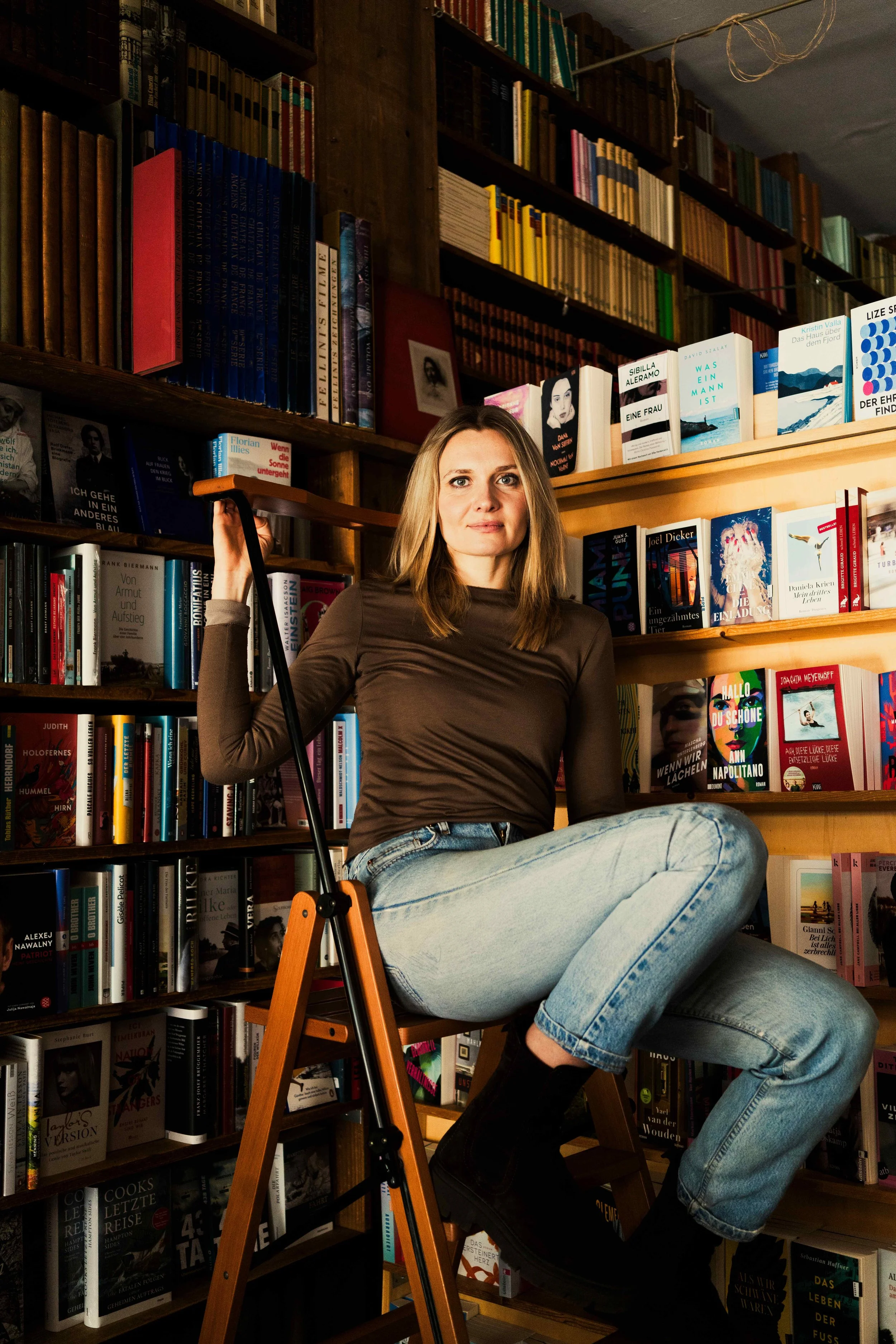Eine junge Frau sitzt auf einer Leiter in einer Buchhandlung, umgeben von ausliegenden Büchern und Bücherregalen, trägt ein braunes langärmeliges Oberteil und Jeans.