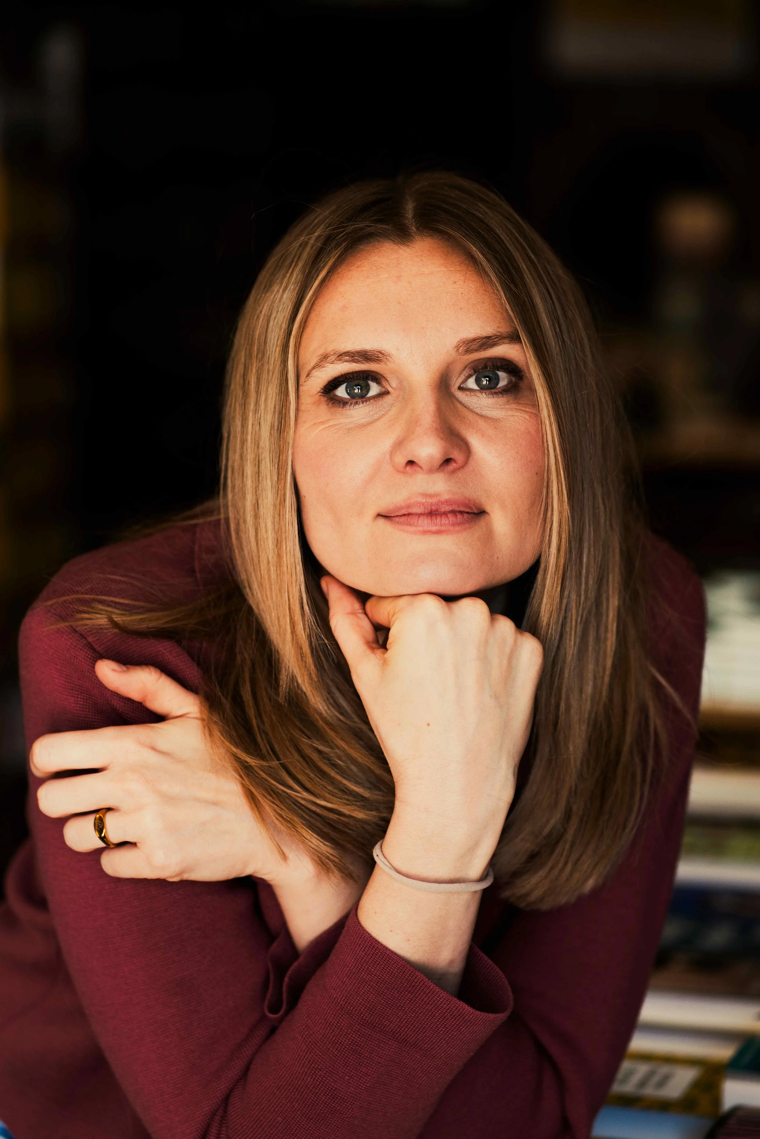 Nahaufnahme einer Frau mit blonden Haaren, blauer Augen und einem dezenten Lächeln, die die Hand unter dem Kinn hält.