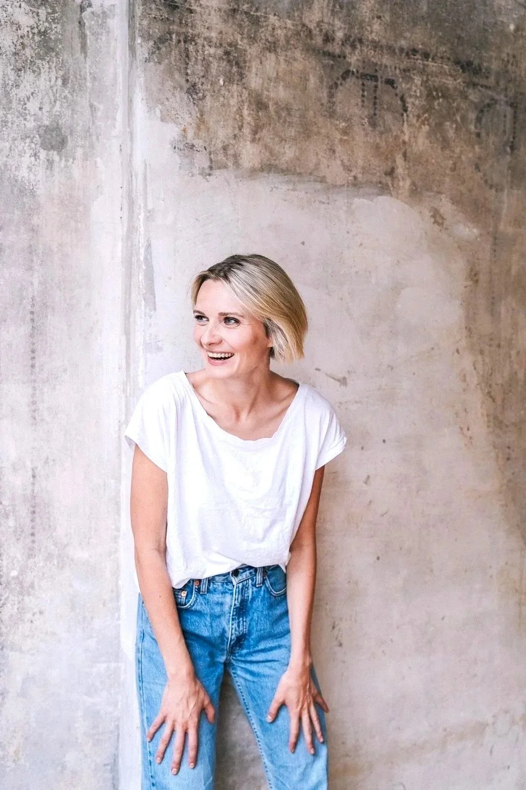 Junge Frau mit blondem Haar trägt ein weißes T-Shirt und blaue Jeans, steht vor einer alten unbehandelten Wand und lächelt.