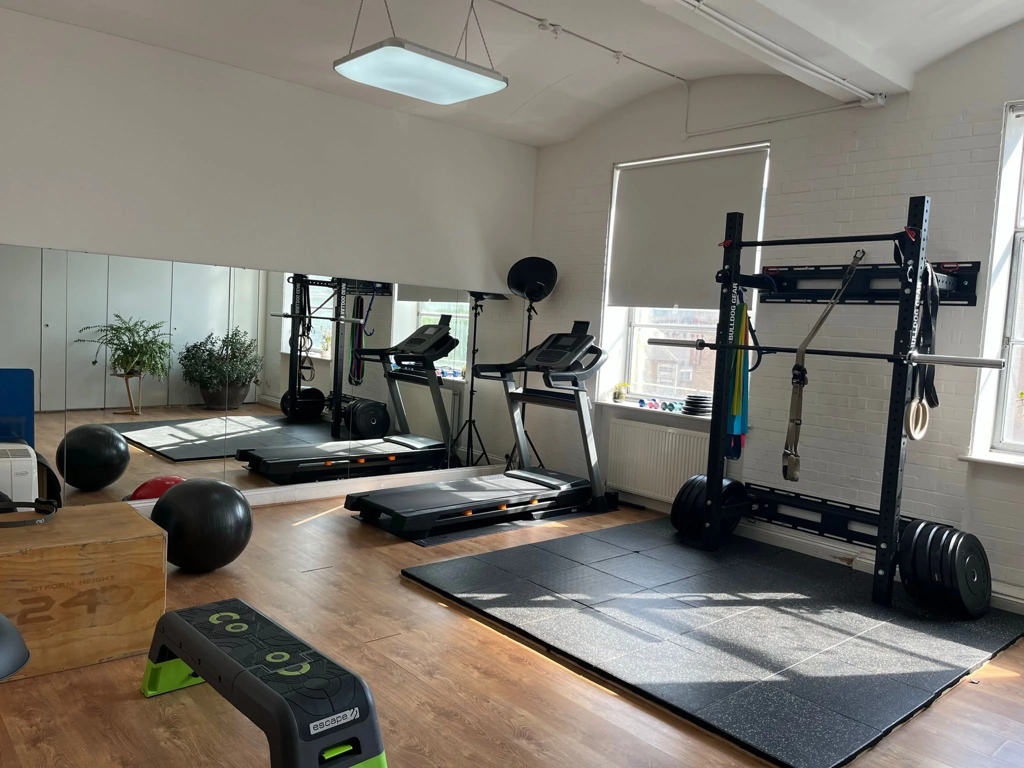 A fitness gym with cardio equipment including treadmills and exercise balls, wooden flooring, large windows, mirrors, and white brick walls.