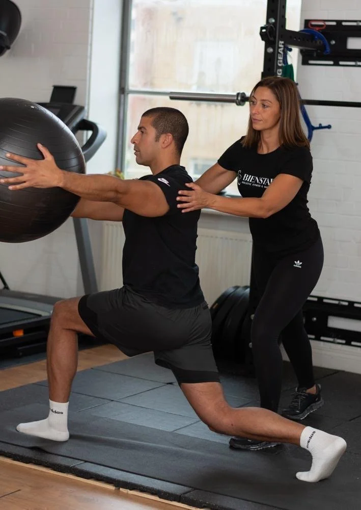 A woman assisting a man in performing a squat exercise with a large exercise ball in a gym.