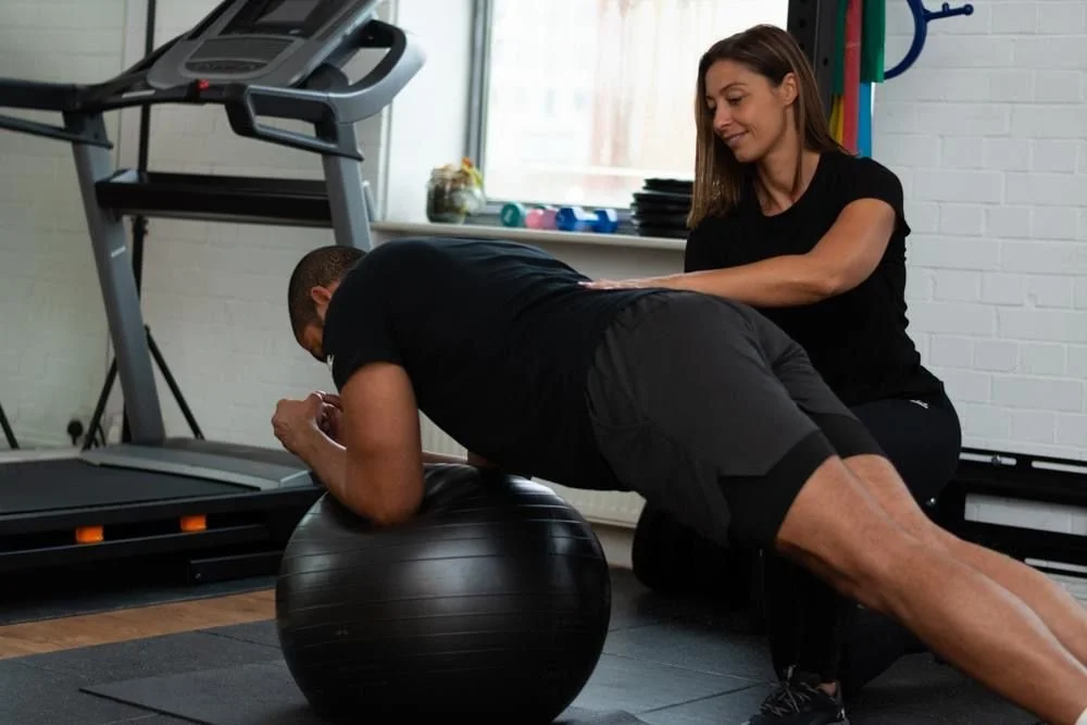 A woman assisting a man performing a plank exercise on a stability ball in a gym.