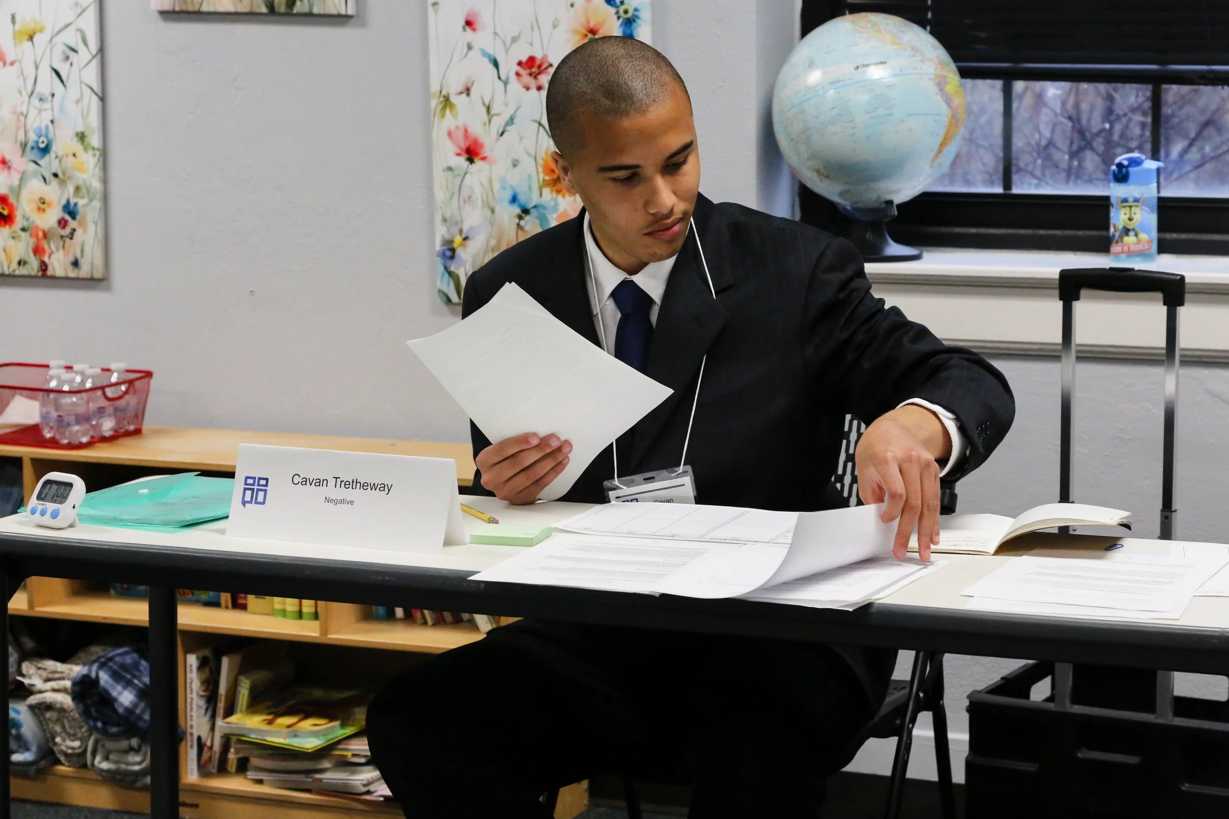 A young student dressed in a black suit, white shirt, and blue tie, sitting at a desk with paperwork, appears to be taking a test or reviewing documents. The student has a short buzz cut hairstyle. There is a nameplate that reads "Cavan Tretheway" with the word "Negative" underneath. The desk also has a timer, green papers, and a calculator. Behind the student, there is a bookshelf, a window with blinds, a globe, and colorful floral curtains.