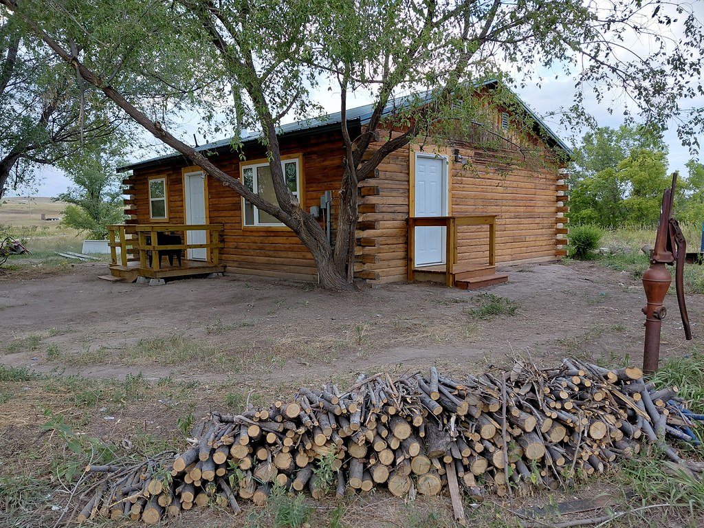 A beautifully renovated wooden log cabin with a small porch and white doors, surrounded by a tree, an open yard, and a pile of firewood in front.