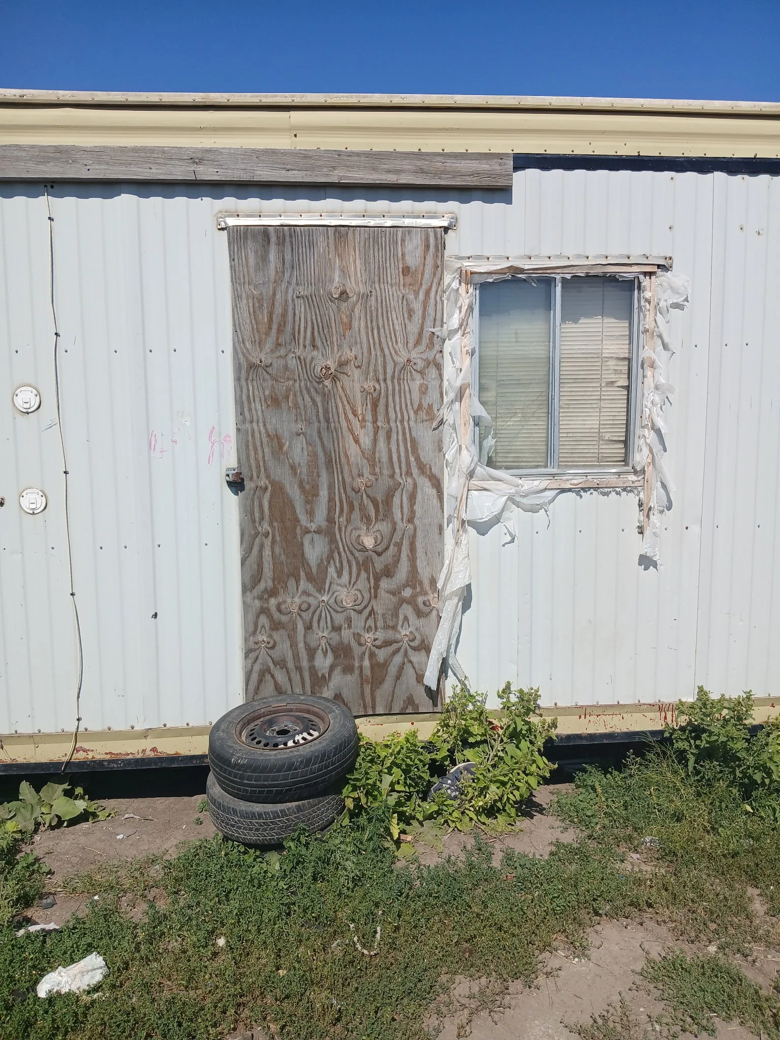 A mobile home with a plywood-covered door and a window surrounded by torn, peeling siding. Two tires are stacked in front, with weeds and grass growing at the base.