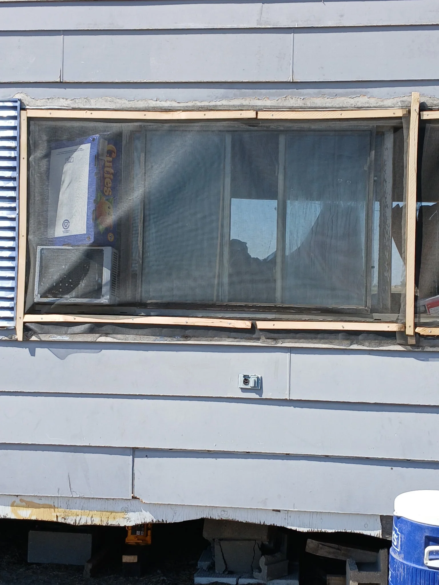 View of a building with a large window, construction framing around the window, and various construction materials including a microwave, a box of Froot Loops cereal, and a bucket. The exterior siding of the building is white with horizontal panels.