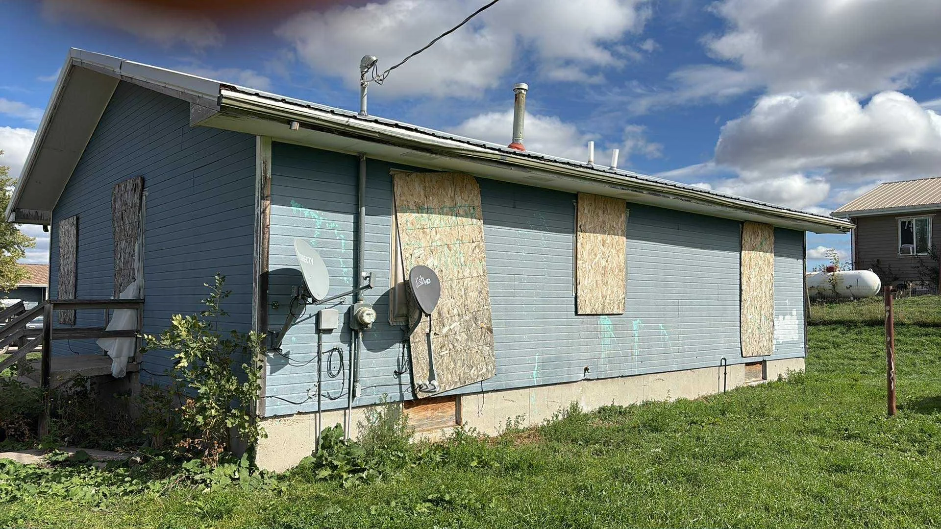 A house with blue siding and boarded-up windows with plywood. Satellite dishes are mounted on the side of the house, and the lawn in front is green with some plants near the foundation. The sky is partly cloudy with blue patches.