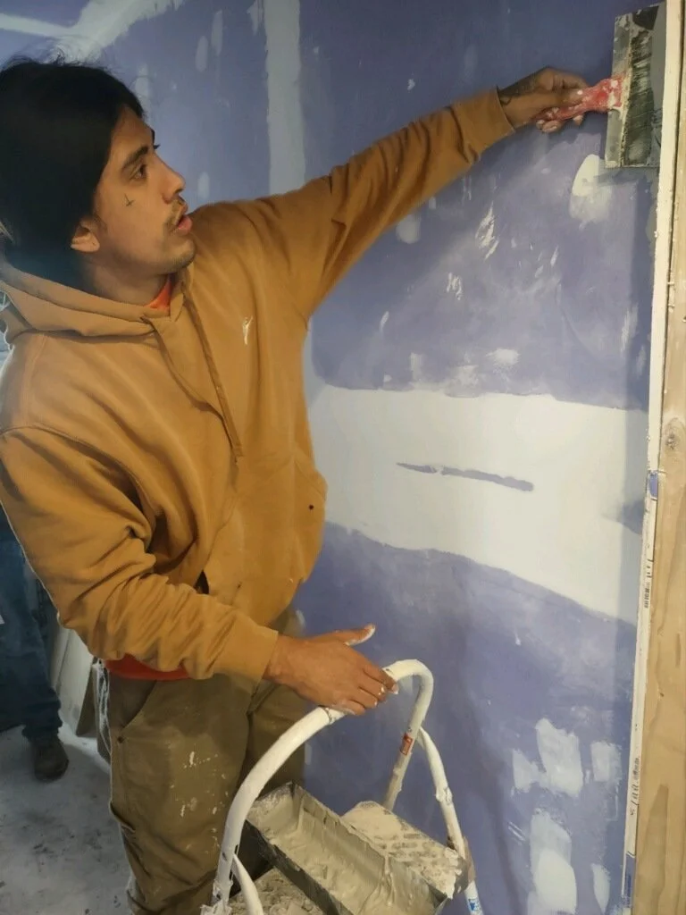 A man in a brown hoodie is painting or preparing a wall for painting with a roller on a ladder.
