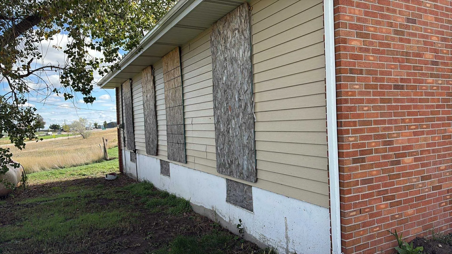 Side of a house with boarded windows, beige siding, and a brick corner, in a rural area with grass and trees.