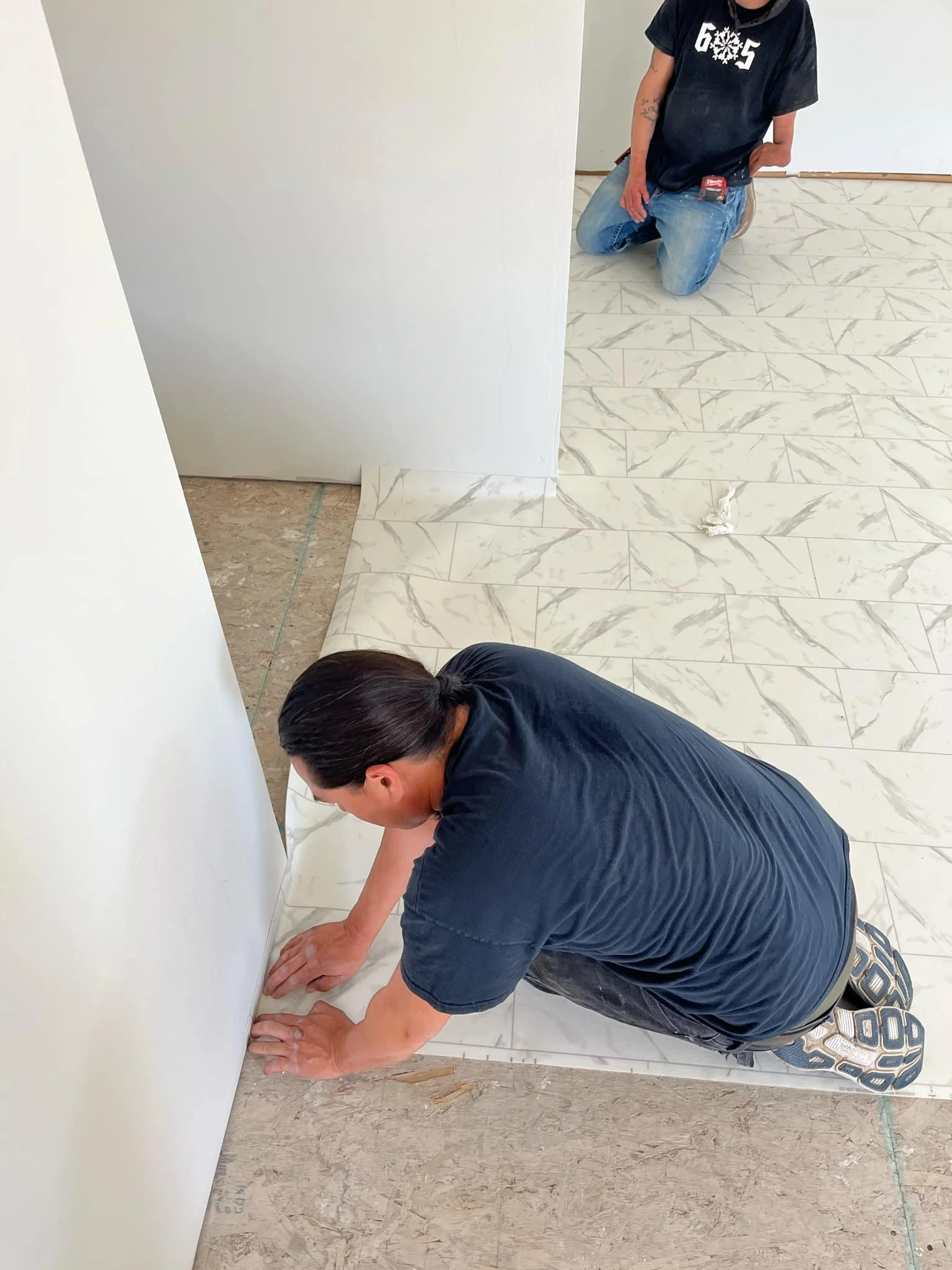 Two workers installing marble-look tiles on the floor, one kneeling and placing tiles, the other kneeling in the background, in a room under construction.