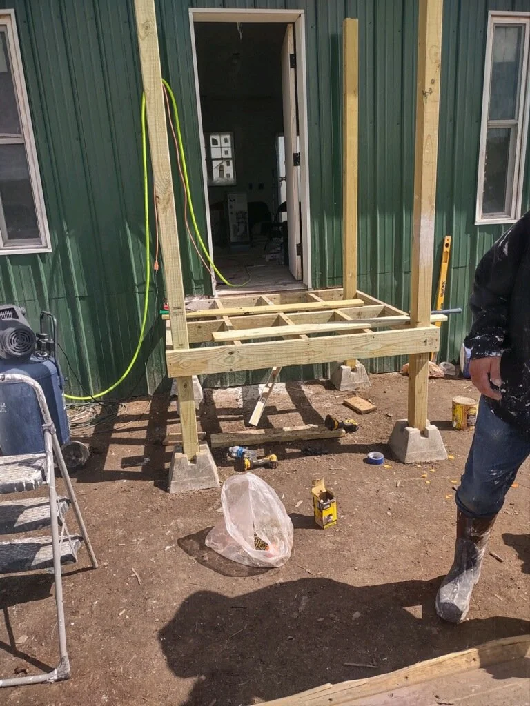 Construction site with a partially built wooden porch in front of a green metal building, tools and materials scattered on the ground, and a person wearing boots and dark clothing on the right side.