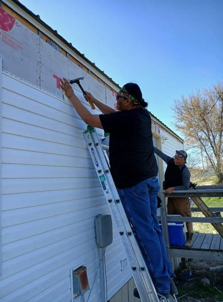 Two men working on siding of a house, one using a hammer on the exterior wall while standing on a ladder, the other standing on a deck observing.