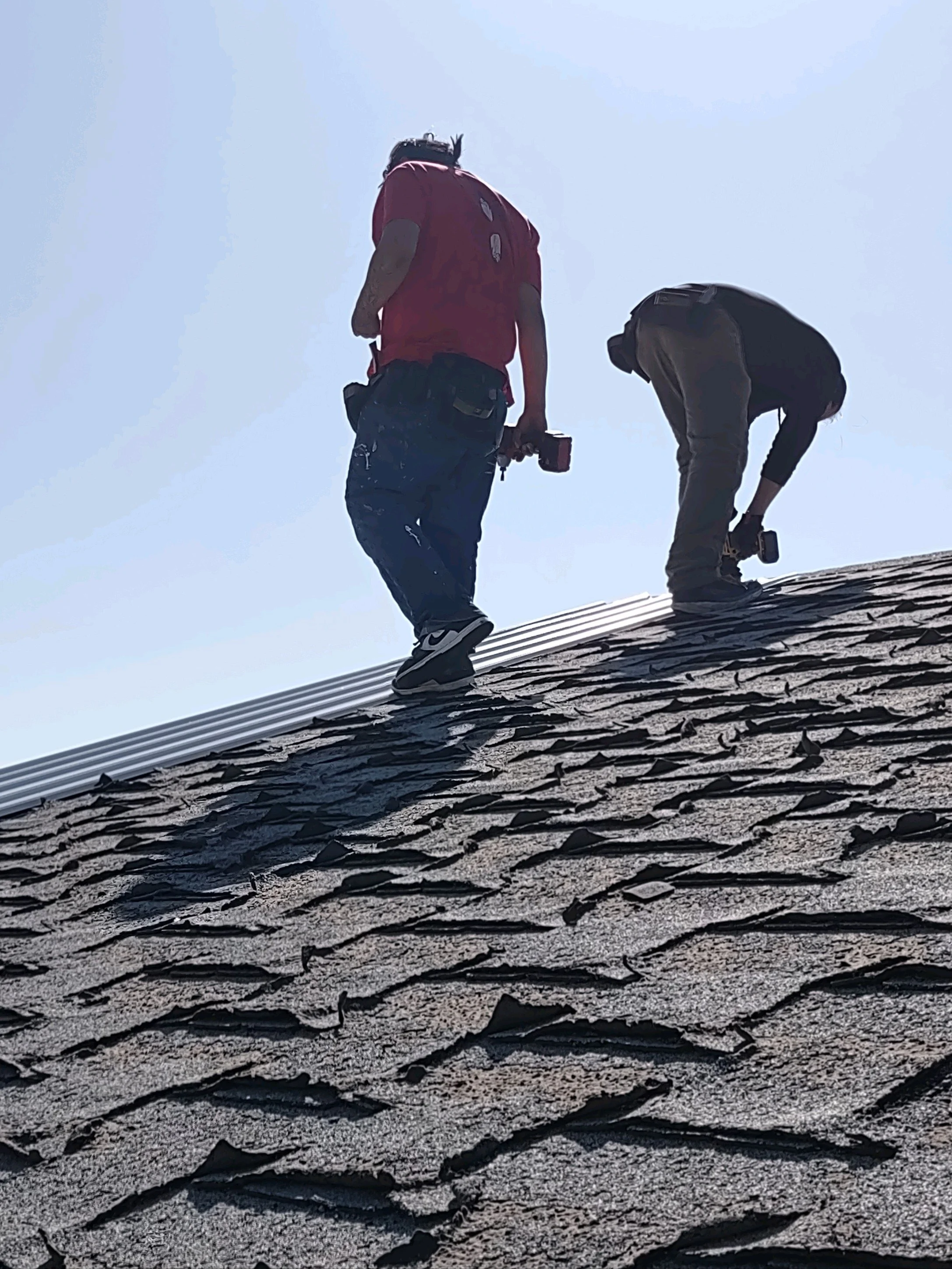 Two workers are installing or repairing roofing shingles on a sloped roof, holding power tools under a clear sky.