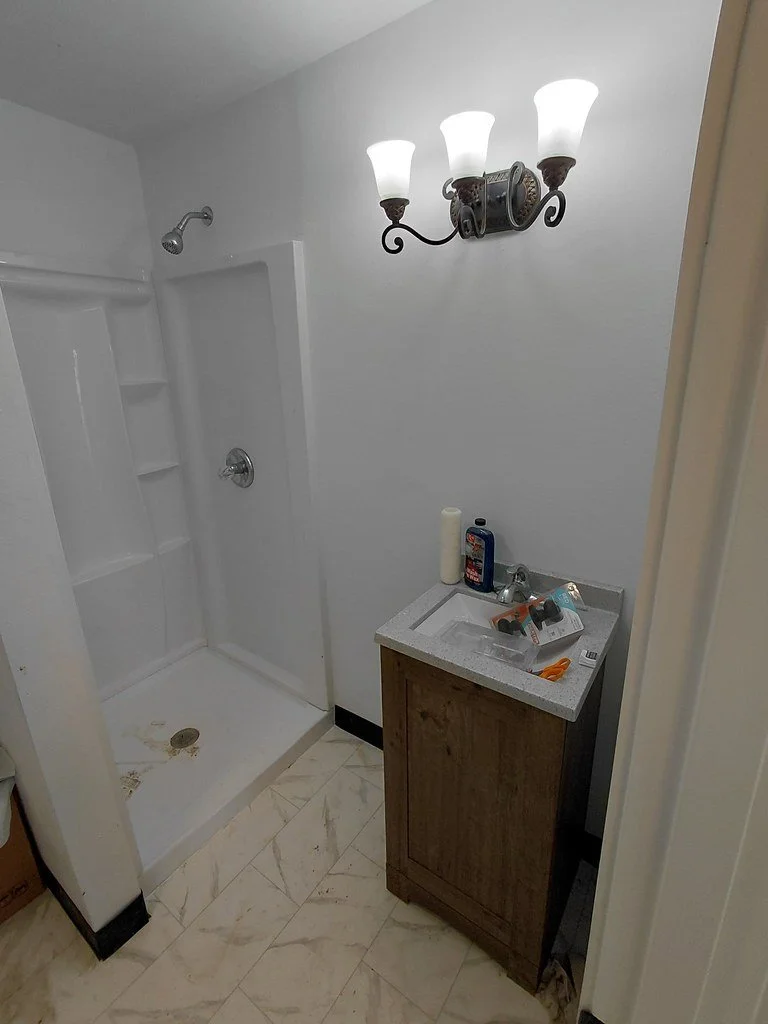 A newly rebuilt small bathroom with a shower enclosure, a wall-mounted light fixture with three frosted glass shades, a brown wooden vanity with a white sink, various toiletries on the counter, and a beige tiled floor.