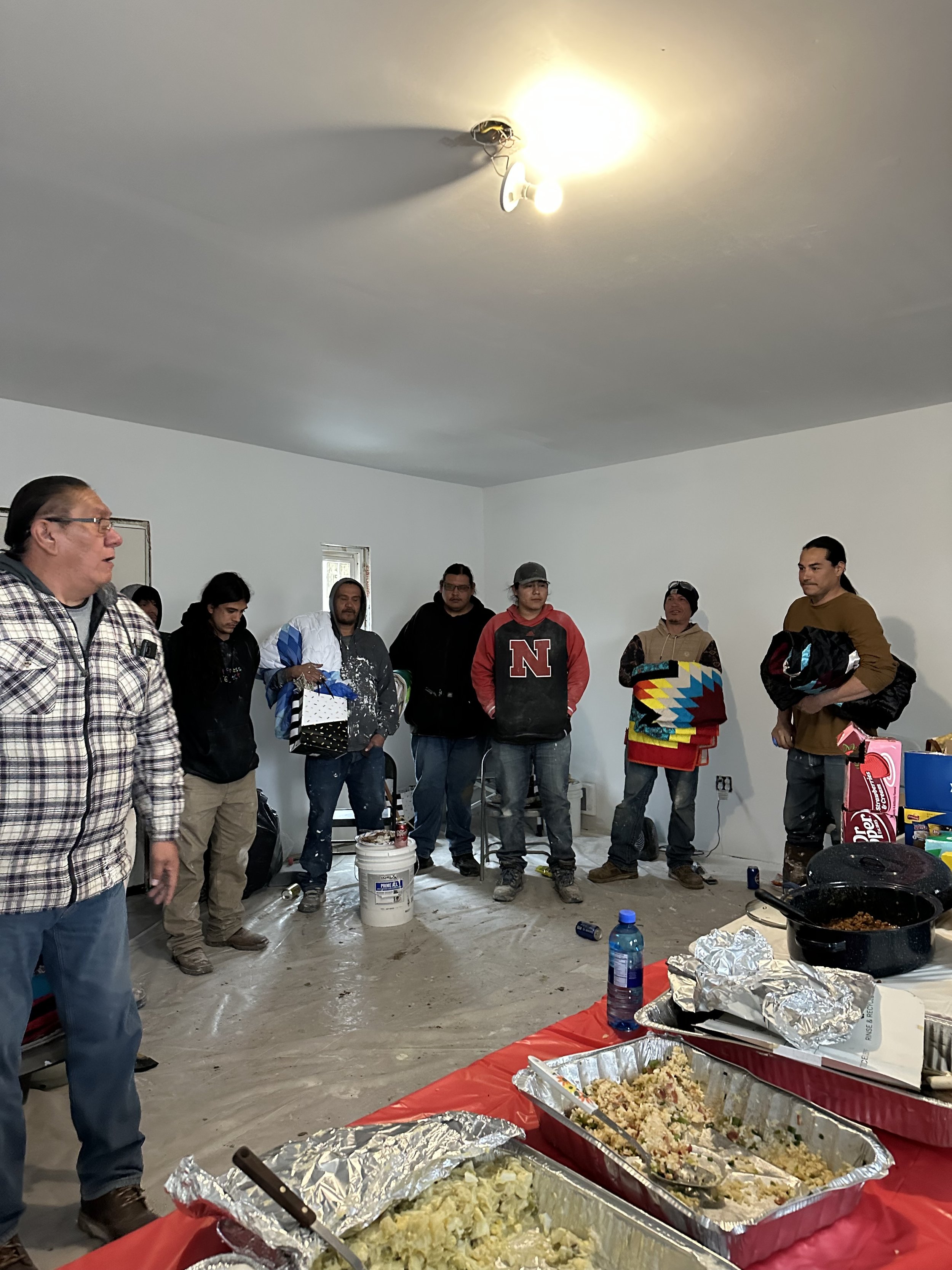 The team is taking a break, standing in a room with a high ceiling and a single light fixture, with tables of food in front of them, including trays of rice, potatoes, and other dishes.