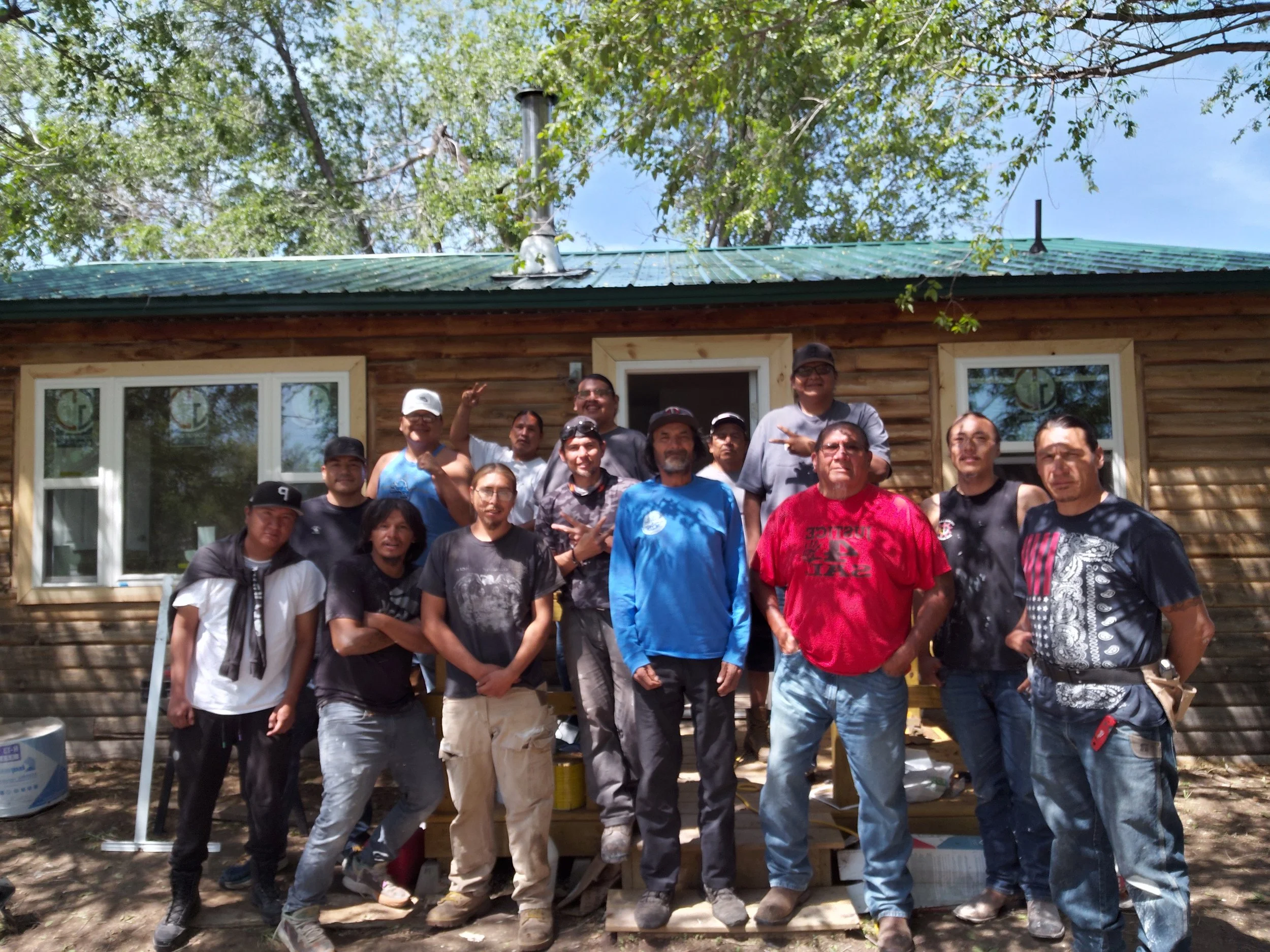 The Lakota Empowerment Group team standing outside a wooden house, under trees, on a sunny day.