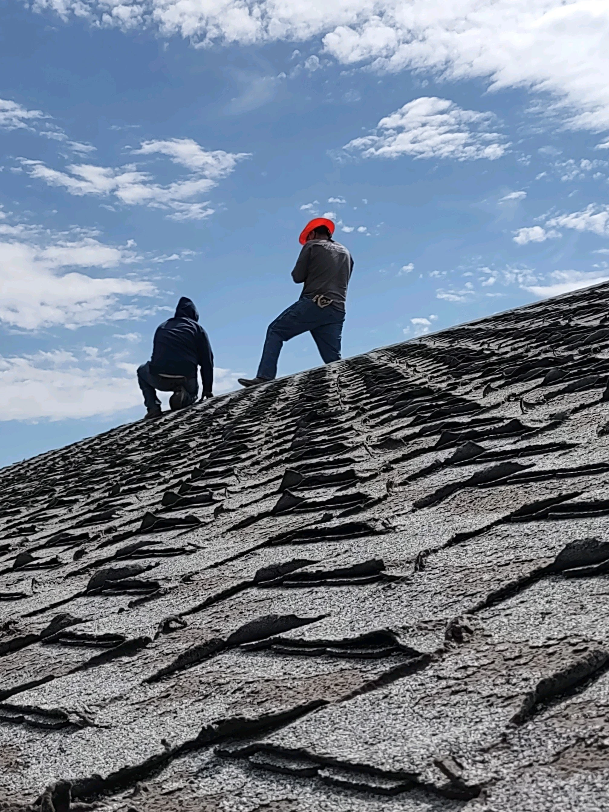 Two individuals on a sloped, textured rooftop under a blue sky with scattered clouds, one wearing a red hat and the other with a hooded jacket.