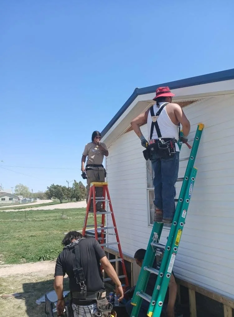 Four workers repairing the exterior of a house, with one person on a step ladder, one on a taller ladder, and two on the ground, during the daytime with a clear blue sky.