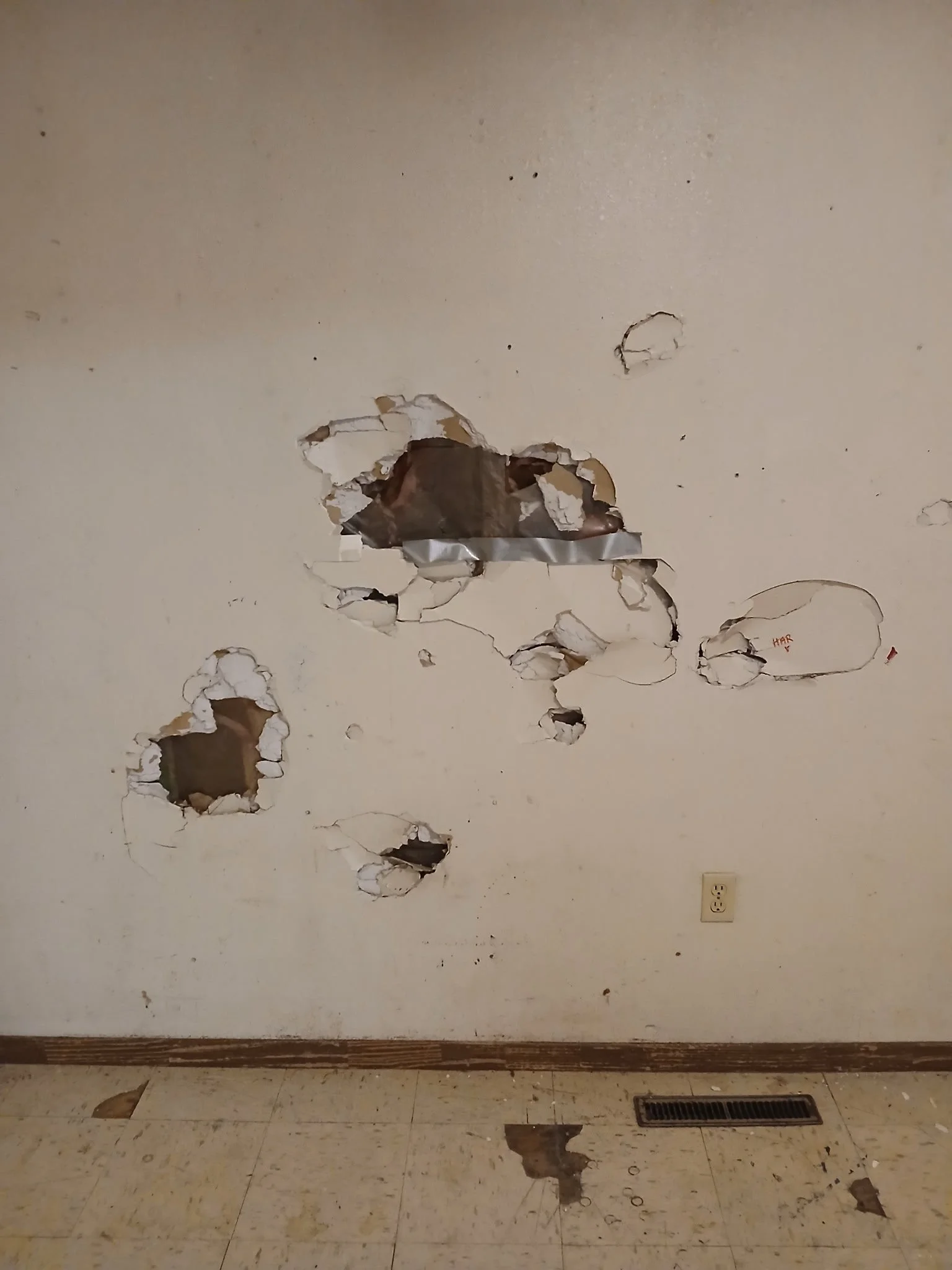 A wall with multiple holes and damage, revealing the wall's interior. The floor has some debris and a floor vent, and there is a power outlet on the wall.