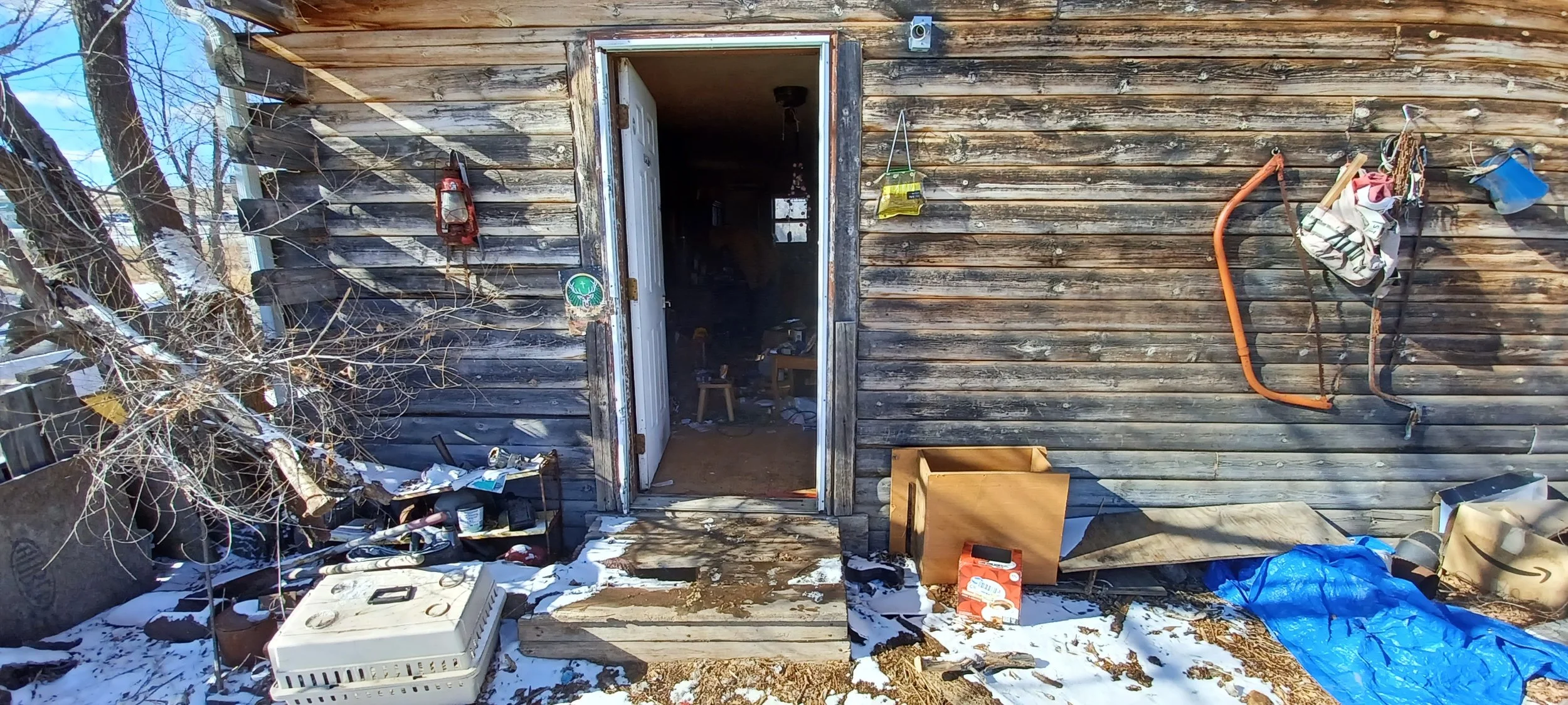 The exterior of a weathered wooden shed with an open door, cluttered with various tools, boxes, and objects outside. Snow is on the ground, and there are garden tools hanging on the wall.