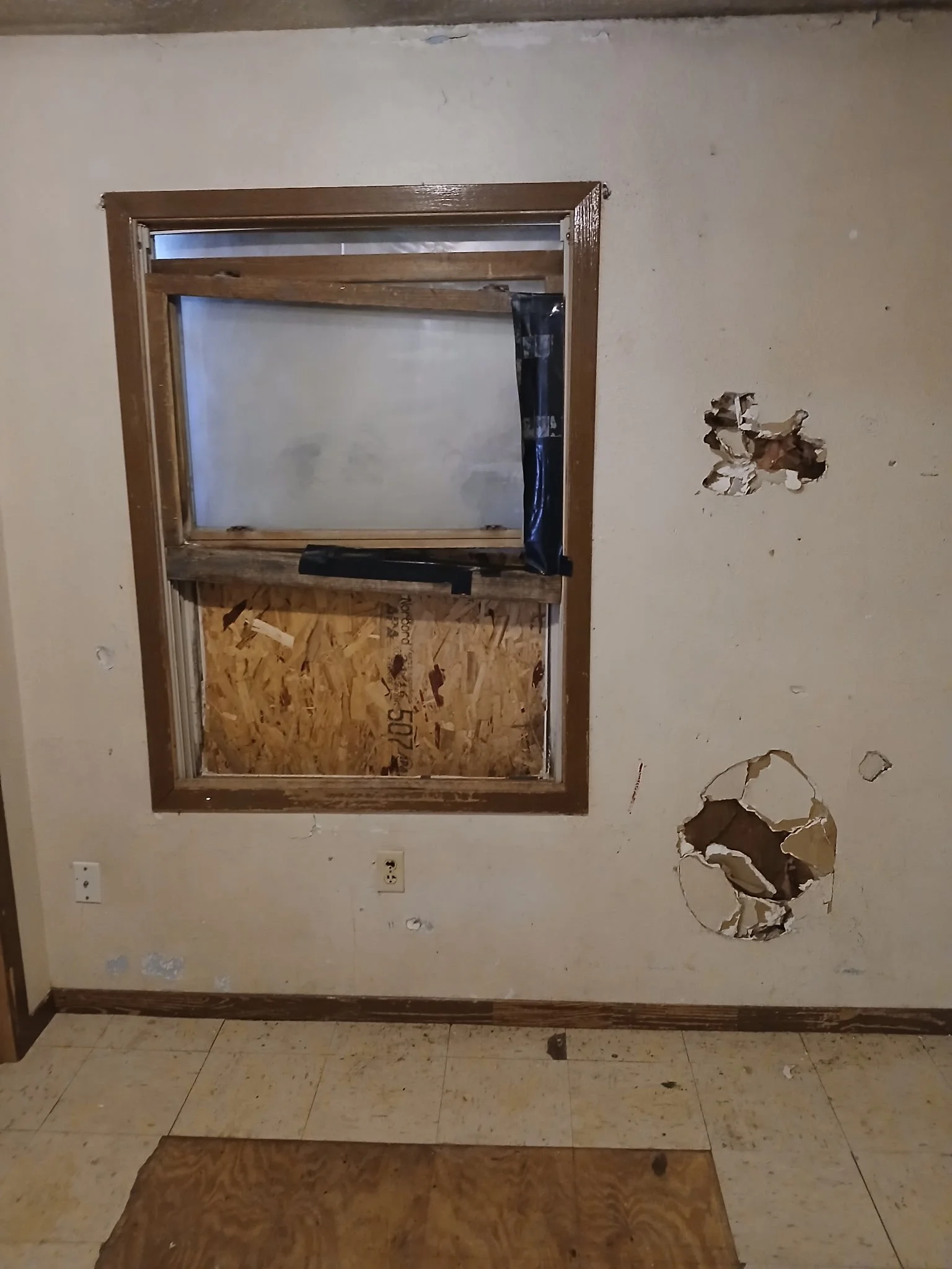 A damaged wall with holes and a window frame with no glass, boarded up with plywood and black tape.