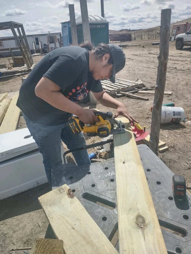 A person wearing a black cap and T-shirt using a yellow power saw to cut a wooden plank, standing outdoors at a construction site with various tools, lumber, and trucks visible in the background.