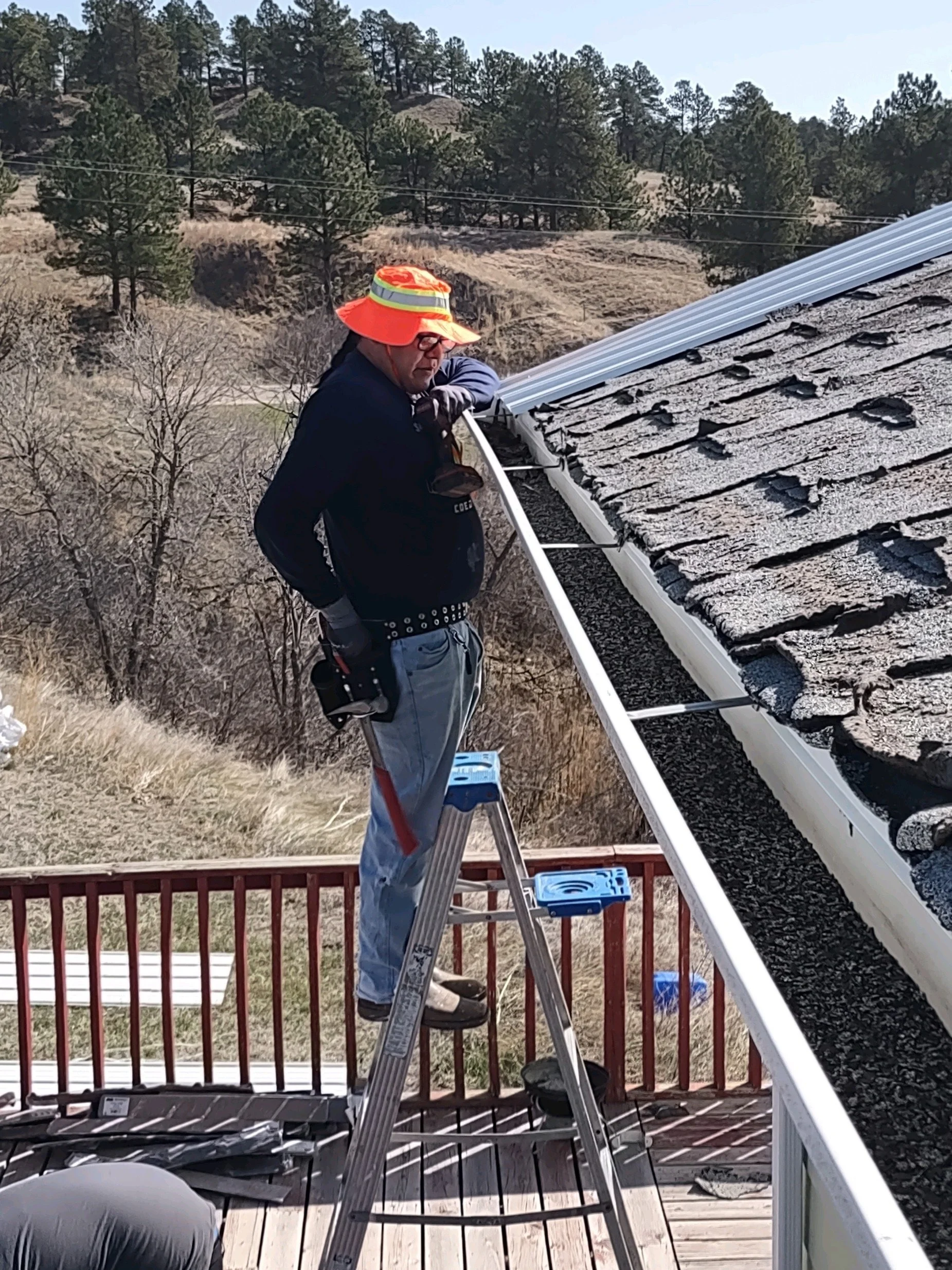 A man wearing a bright orange hat and sunglasses working on roofing shingles on a house's roof, standing on a blue step stool, with a tool belt and gloves, in a rural area with trees and hills in the background.