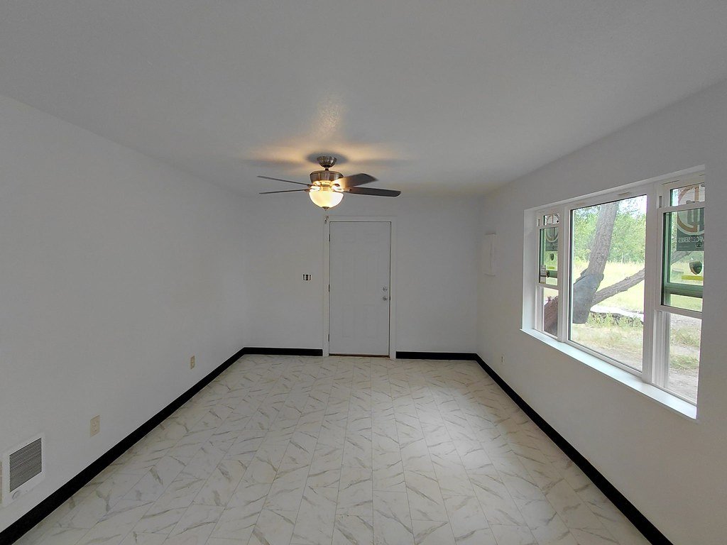 A clean empty room with white walls, black baseboards, large window showing a tree outside, and a ceiling fan with a light in the center of the ceiling.
