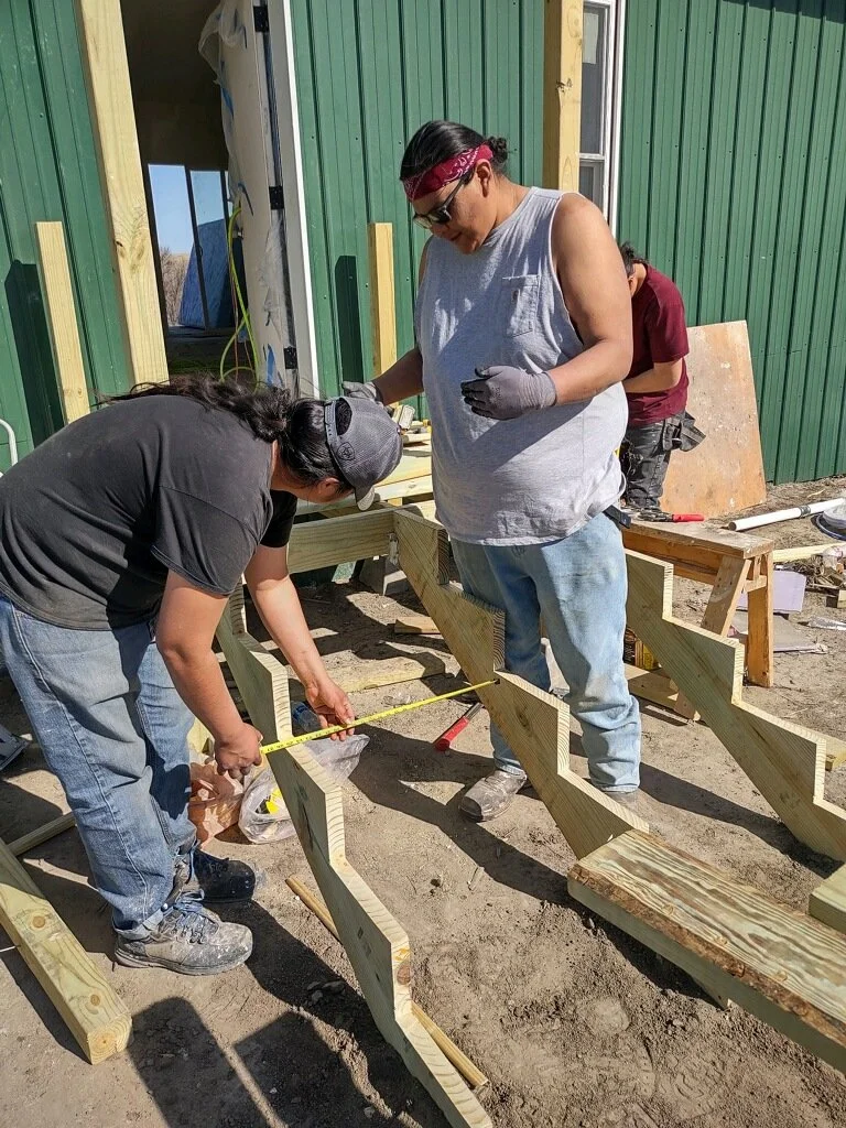 Two women are building a wooden structure outside a green building, measuring and assembling the wood on the ground.