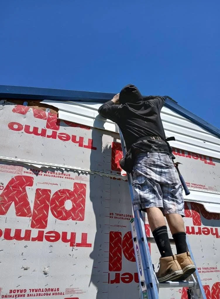 A person wearing a black hoodie, plaid shorts, and brown work boots standing on a ladder, installing or inspecting siding on a building under a clear blue sky.