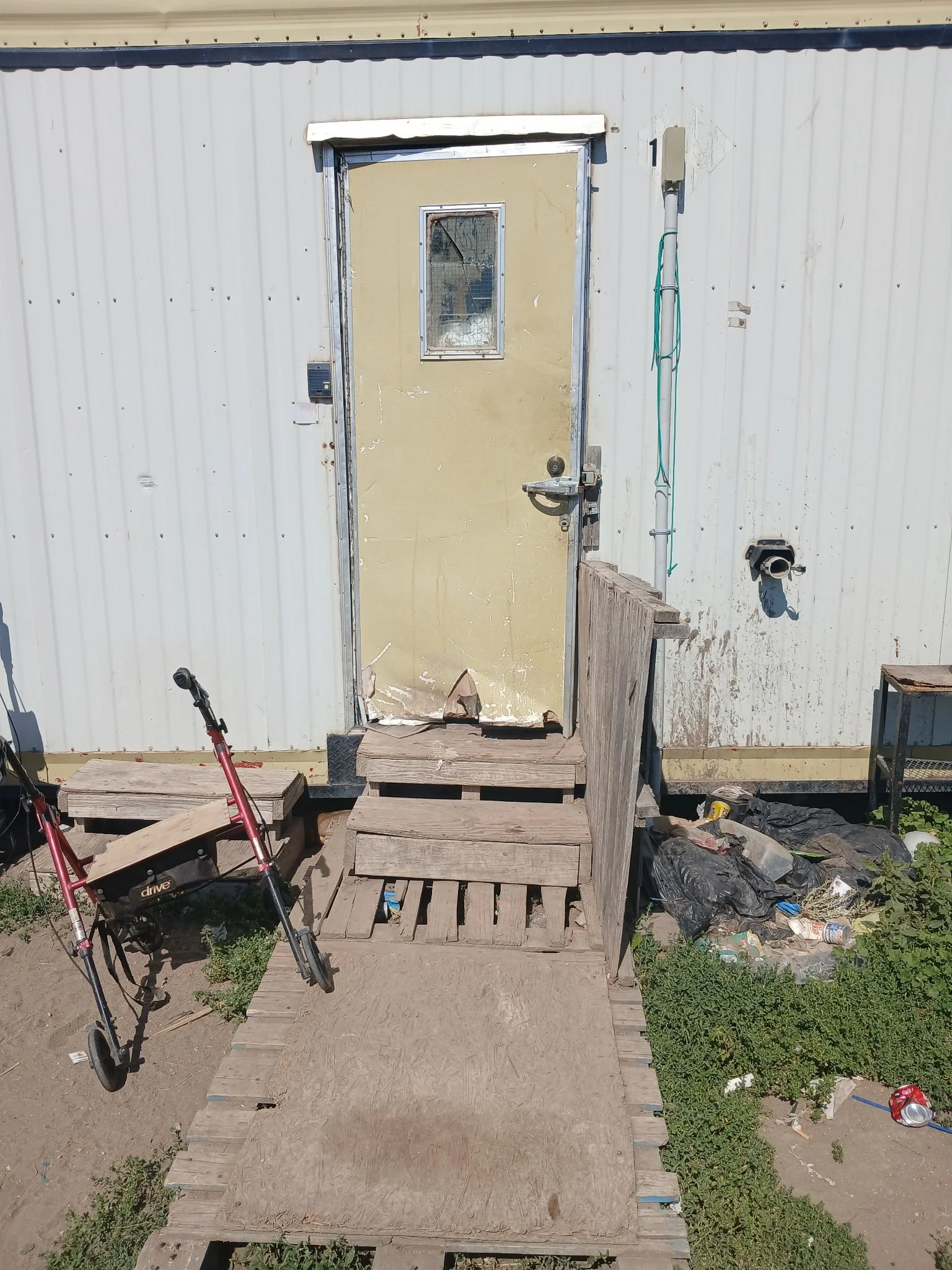 A metal door with a small window is set in a corrugated metal building. The door has a damaged bottom and a handle with an additional lock. There is a wooden ramp and steps leading up to the door. To the left, there is a red mobility scooter. Debris,