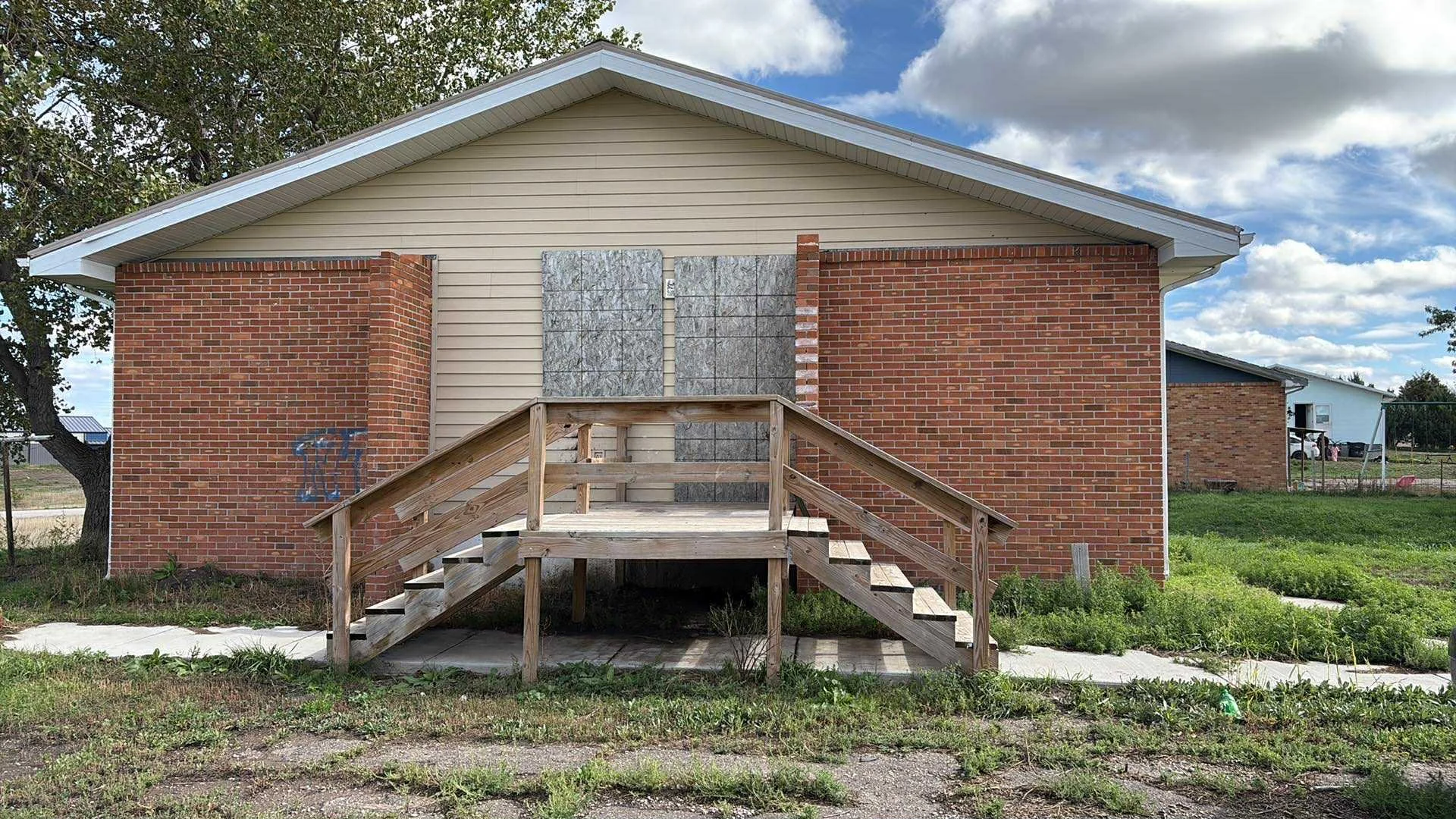 Back of house with boarded-up windows, plywood covering windows, brick and siding exterior, wooden stairs leading to door, overgrown grass, and a sidewalk in a suburban area.