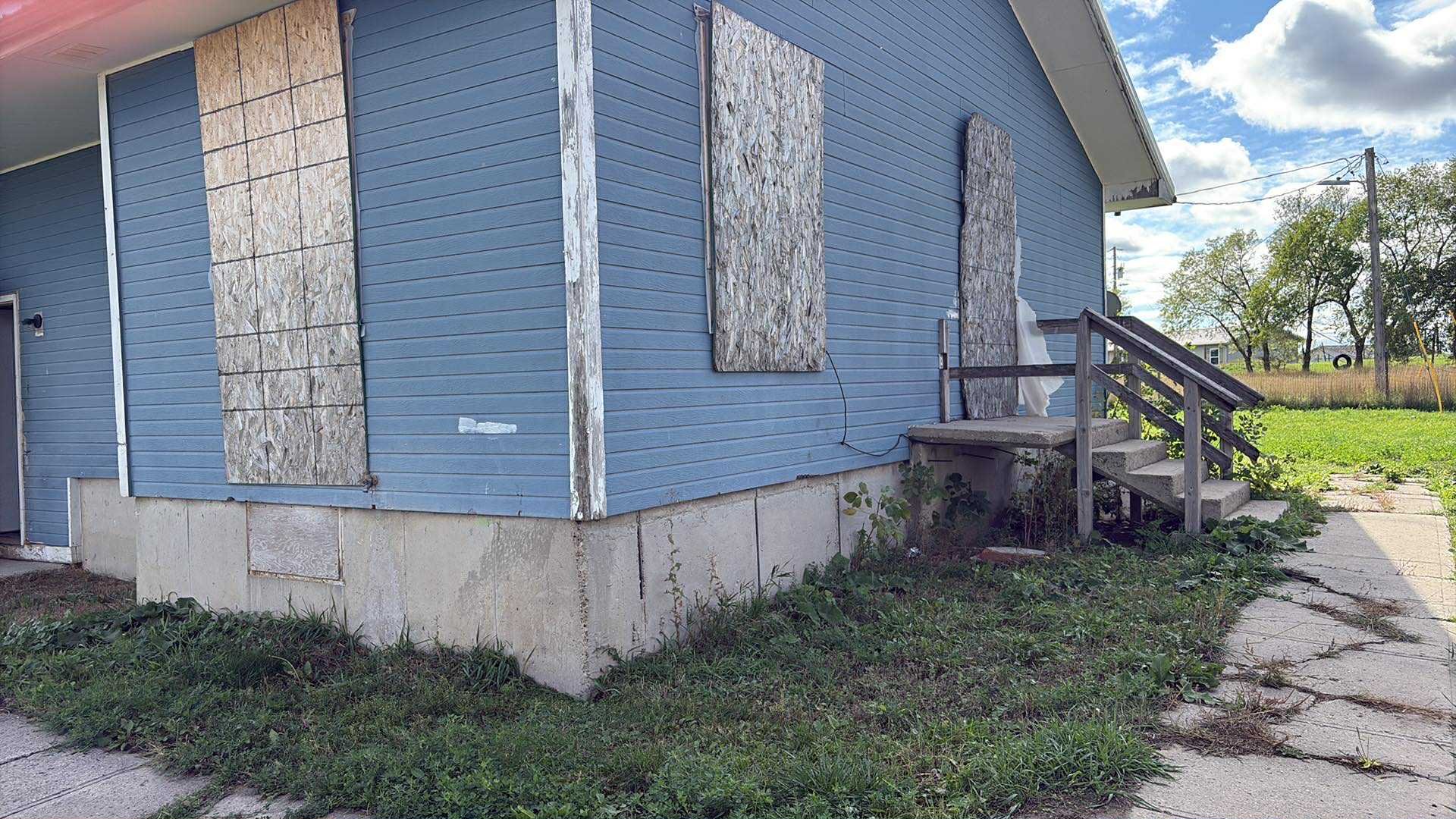 Blue house with boarded-up windows and front steps in a rural area, under a partly cloudy sky.