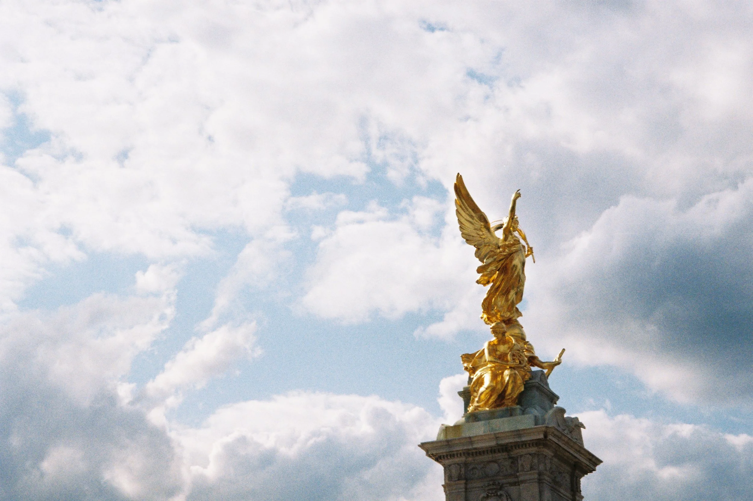 A golden statue of a winged woman holding a staff, atop a stone pedestal, against a backdrop of cloudy sky.